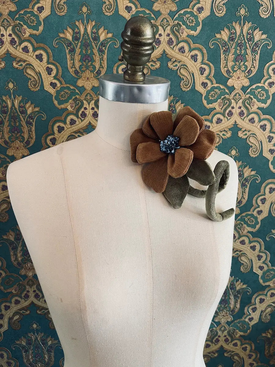 Broochella Velluto brown and  green handmade velvet flower designer brooch with stem and leaves. Styled on a mannequin. The background is green and gold damask wallpaper