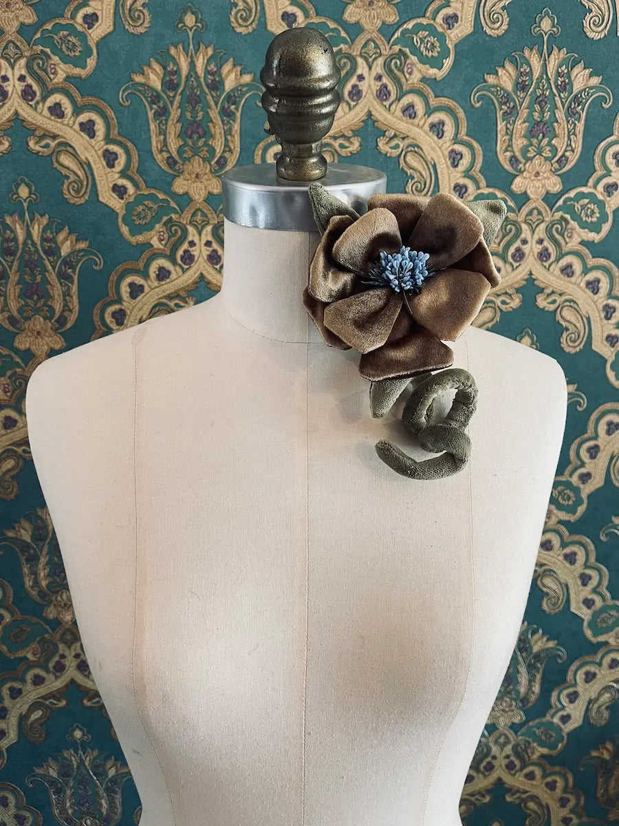 Broochella Velluto gold and  green handmade velvet flower designer brooch with stem and leaves. Styled on a mannequin. The background is green and gold damask wallpaper