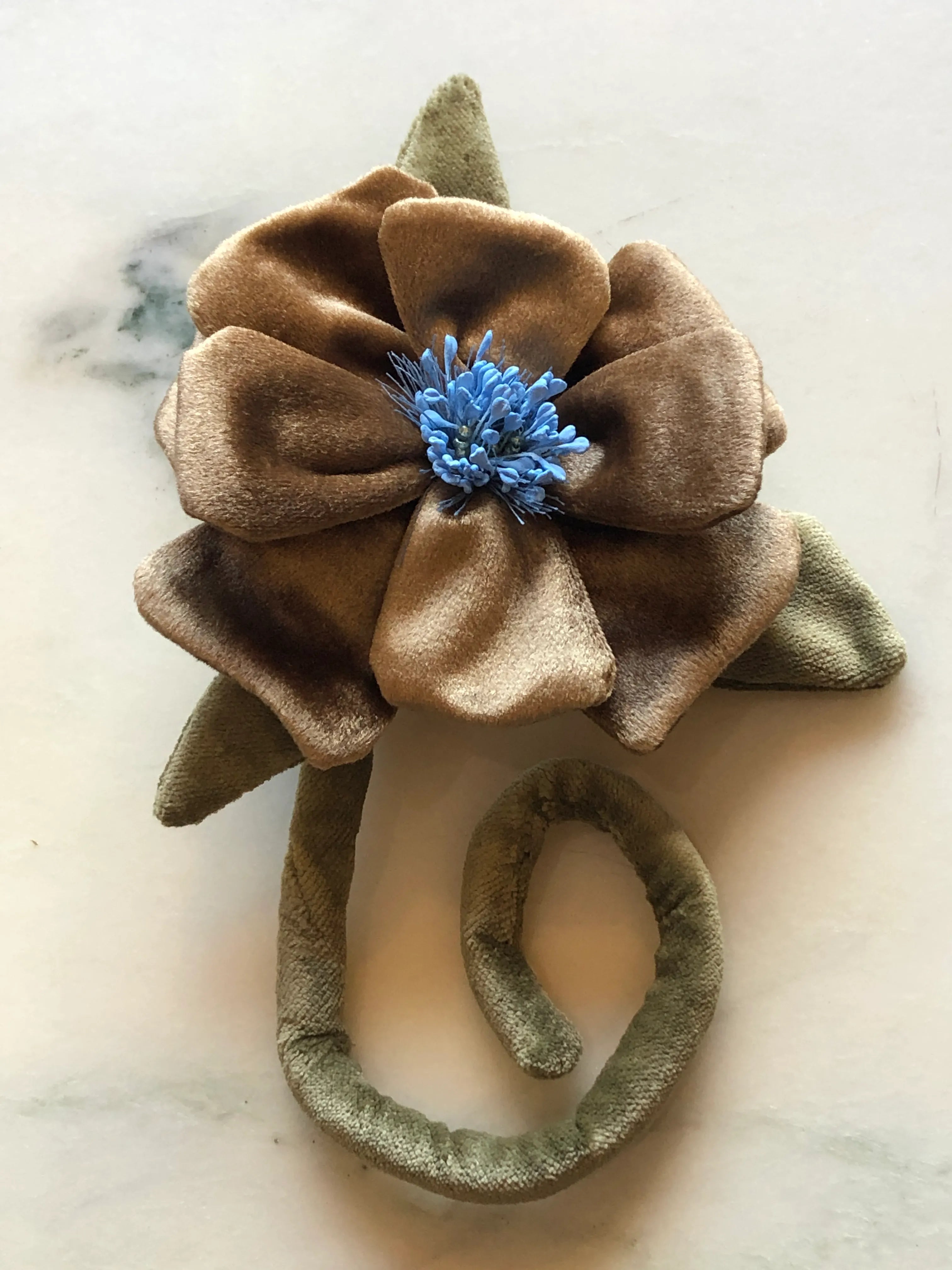 A Broochella handmade fabric flower brooch featuring a large gold velvet flower with a blue velvet center, olive green leaves, and a matching olive green elastic band, displayed on a light marble surface.