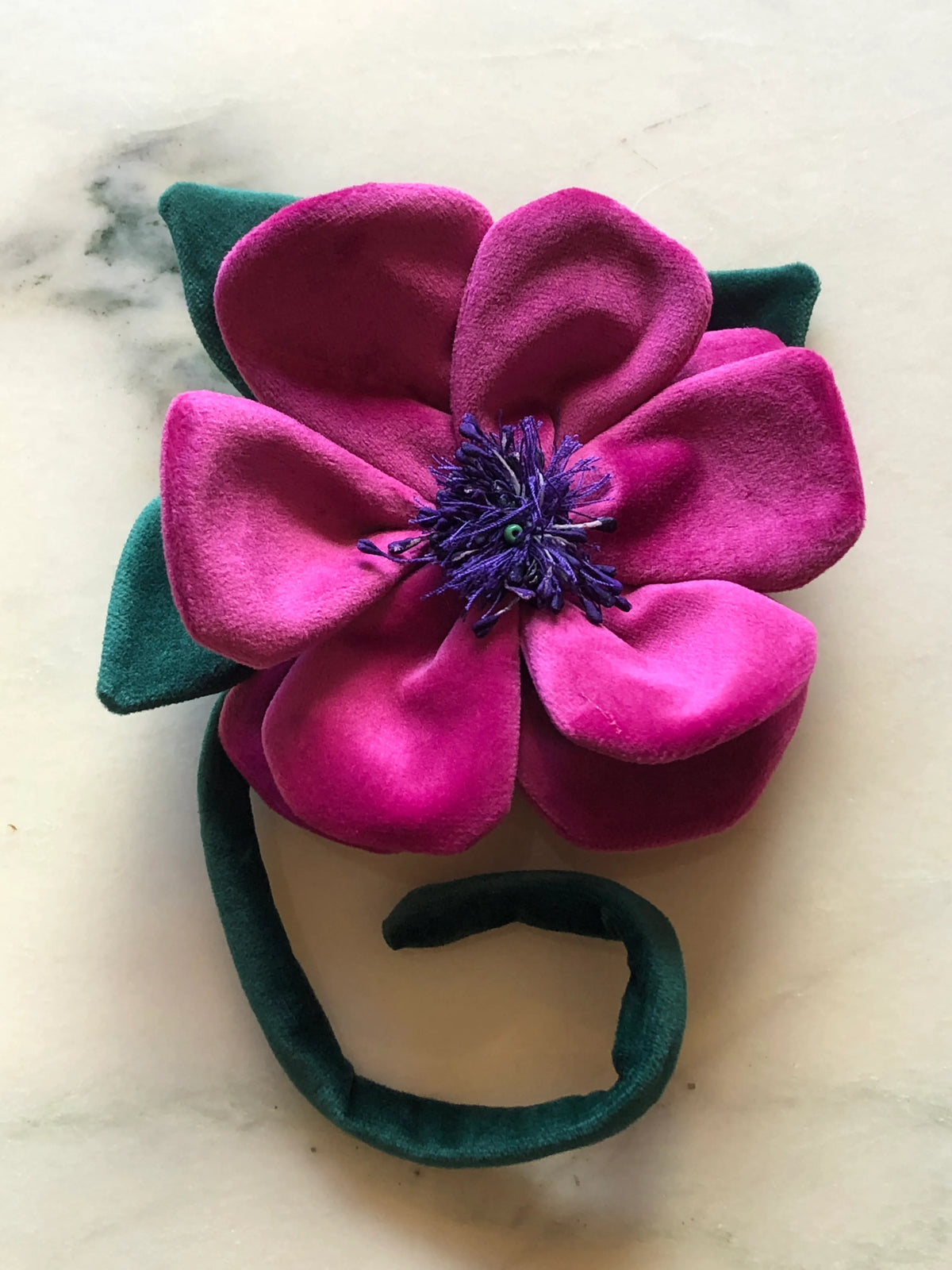 A Broochella Velluto handmade velvet flower brooch featuring a large magenta-pink flower with dark green leaves and a purple centre, attached to a matching dark green velvet headband, displayed on a light marble surface.