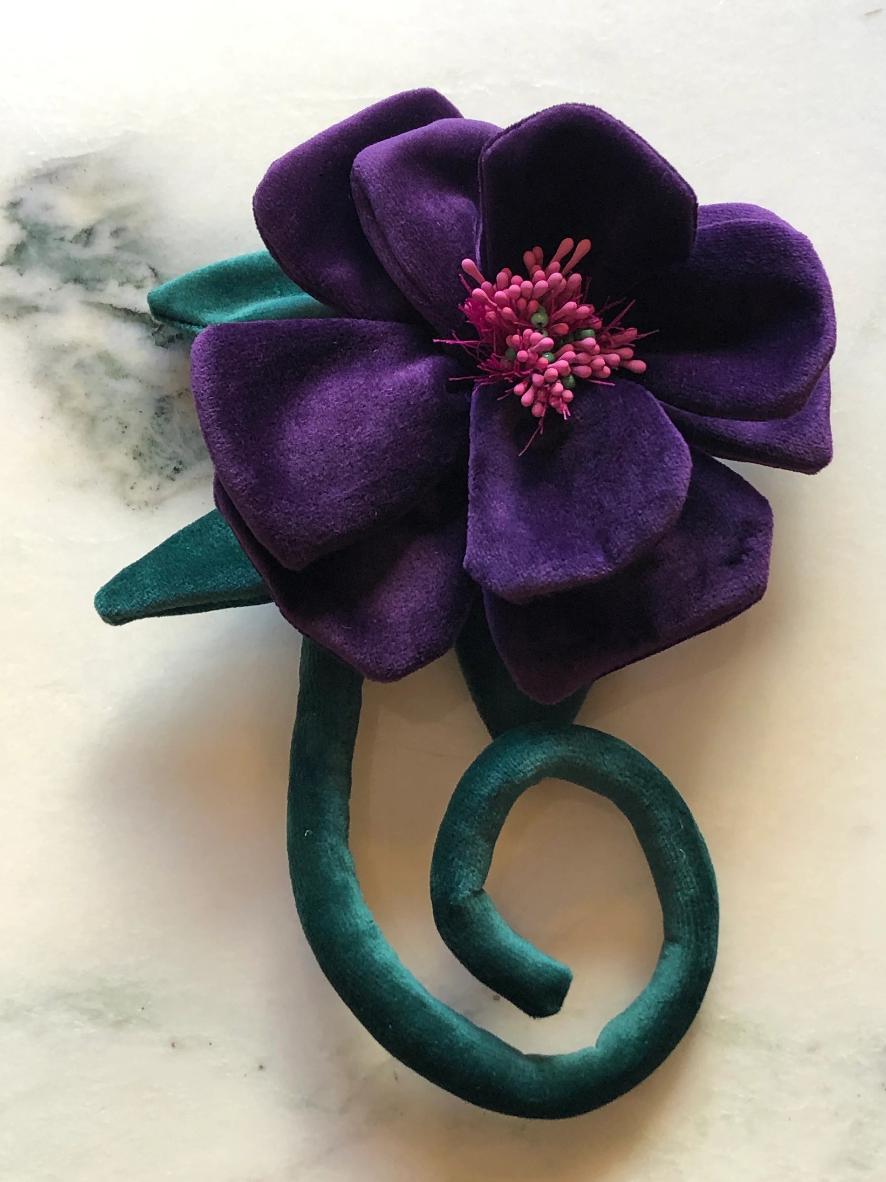 A  Broochella Velluto purple velvet flower brooch with a pink stamen center and a twisted teal velvet stem, displayed on a light marble surface.