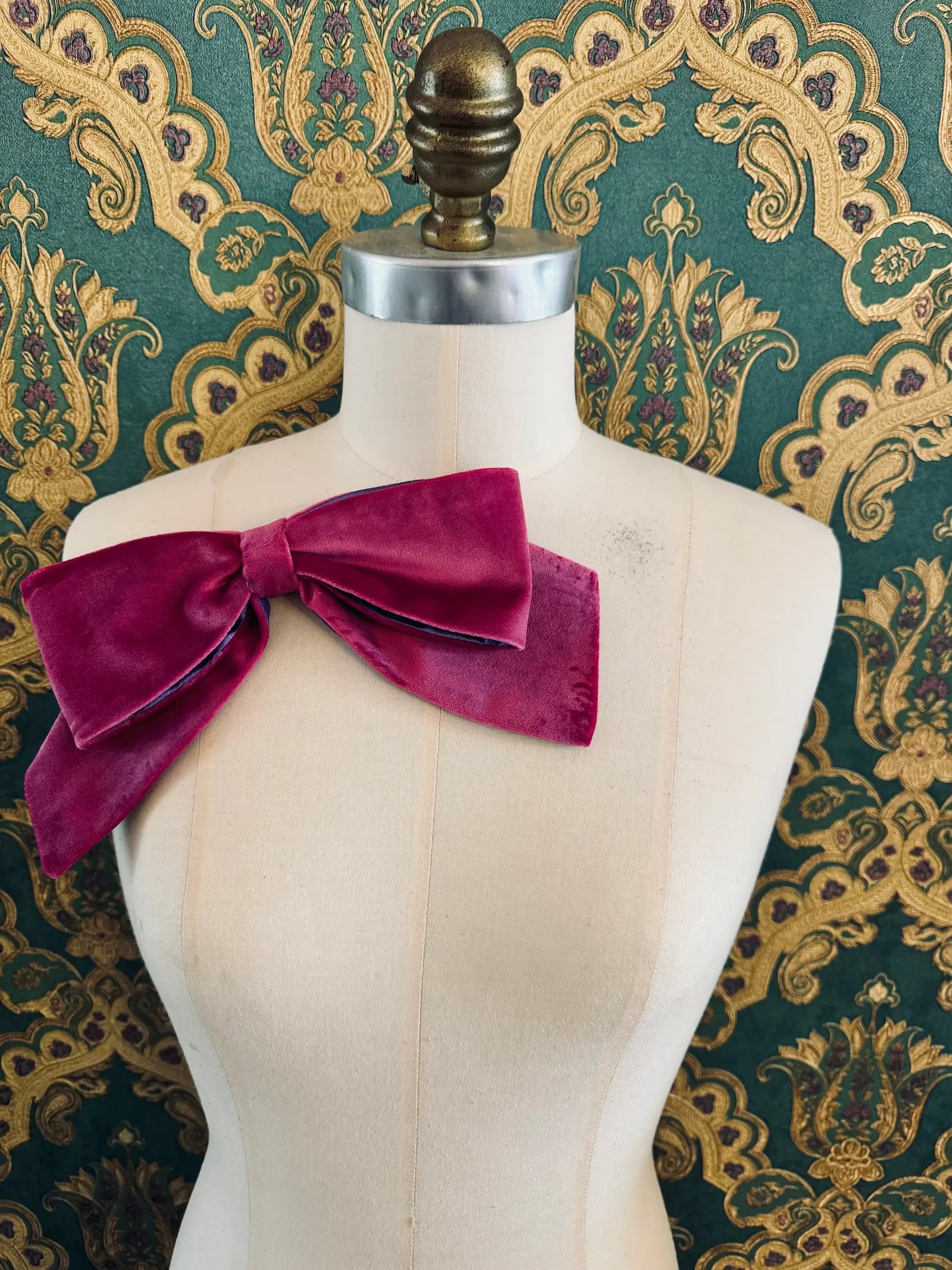 Broochella large velvet handmade bow brooch in magenta and purple. Pictured on a mannequin.  The background is green and gold damask wallpaper.