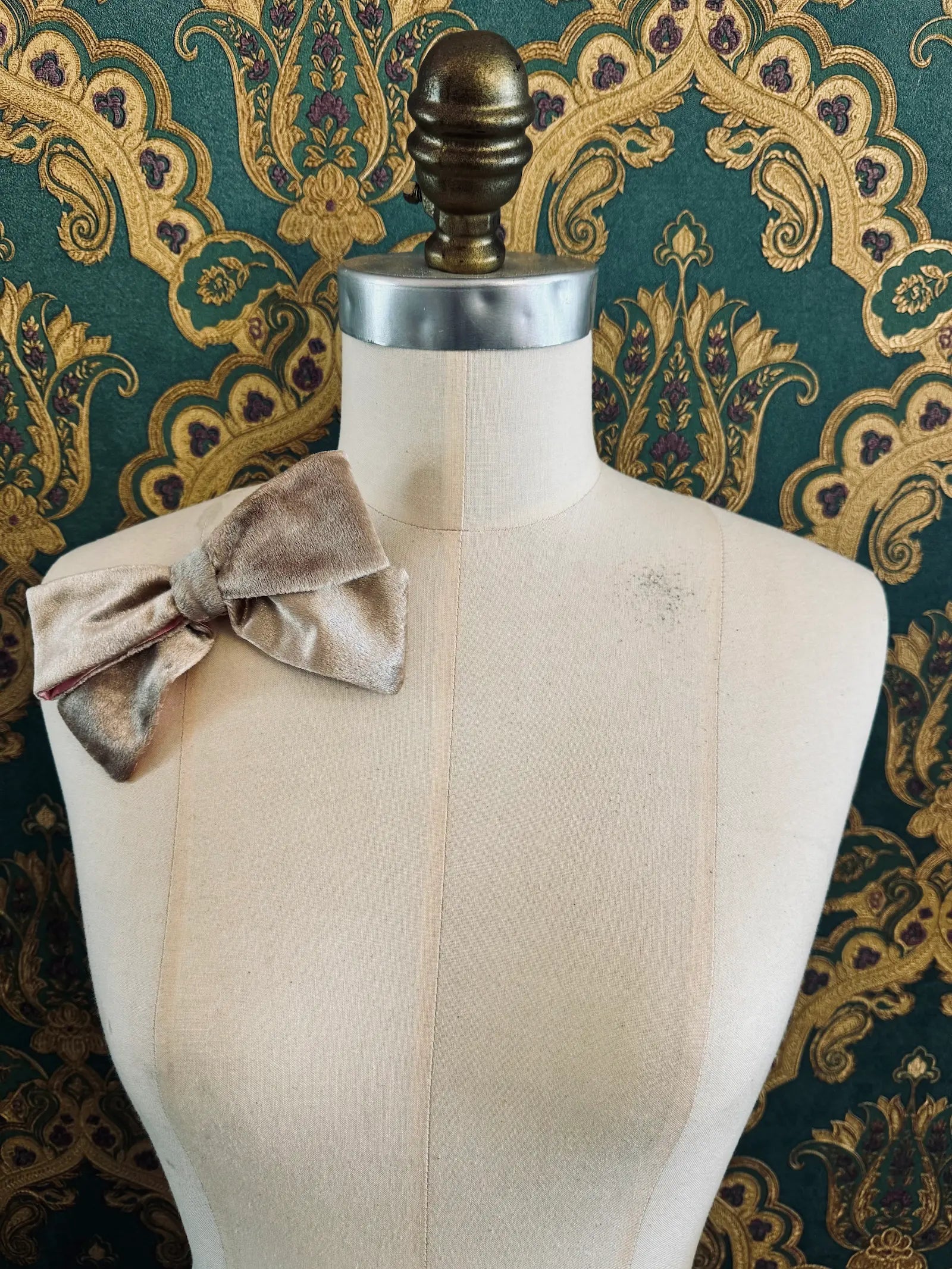 Broochella small velvet handmade bow brooch in champagne and pink. Pictured on a mannequin.  The background is green and gold damask wallpaper.