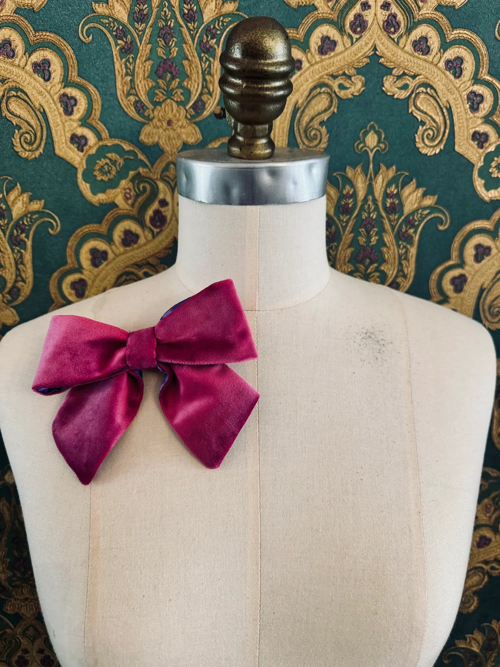 Broochella small velvet handmade bow brooch in magenta and purple. Pictured on a mannequin.  The background is green and gold damask wallpaper.