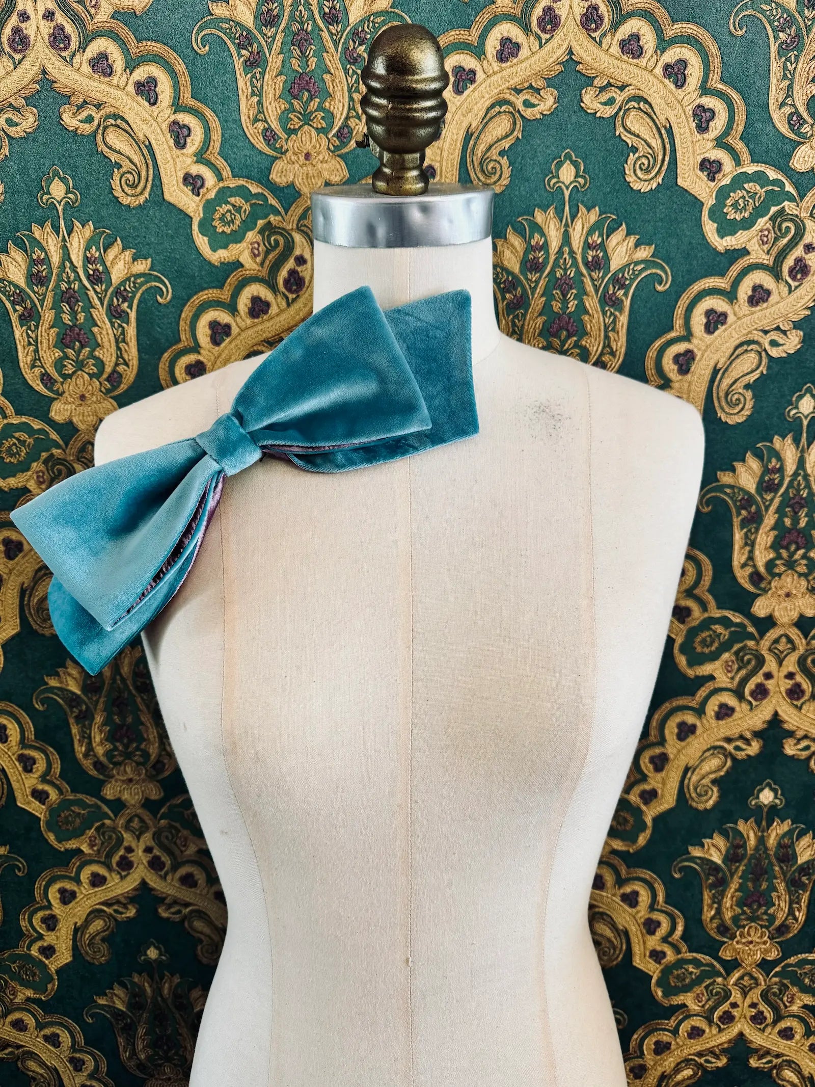 Broochella large velvet handmade bow brooch in aqua and purple. Pictured on a mannequin.  The background is green and gold damask wallpaper.