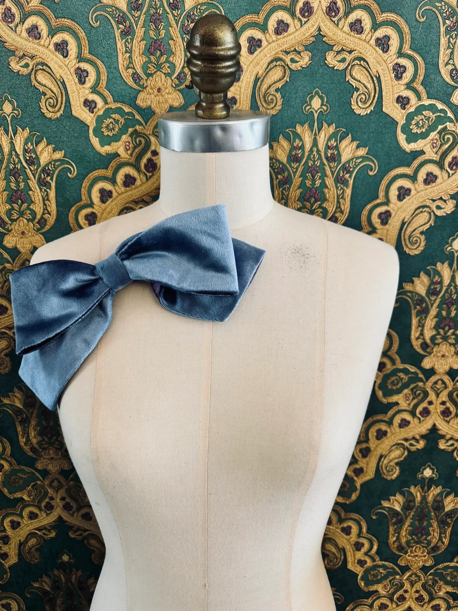 Broochella large velvet handmade bow brooch in blue and purple. Pictured on a mannequin.  The background is green and gold damask wallpaper.