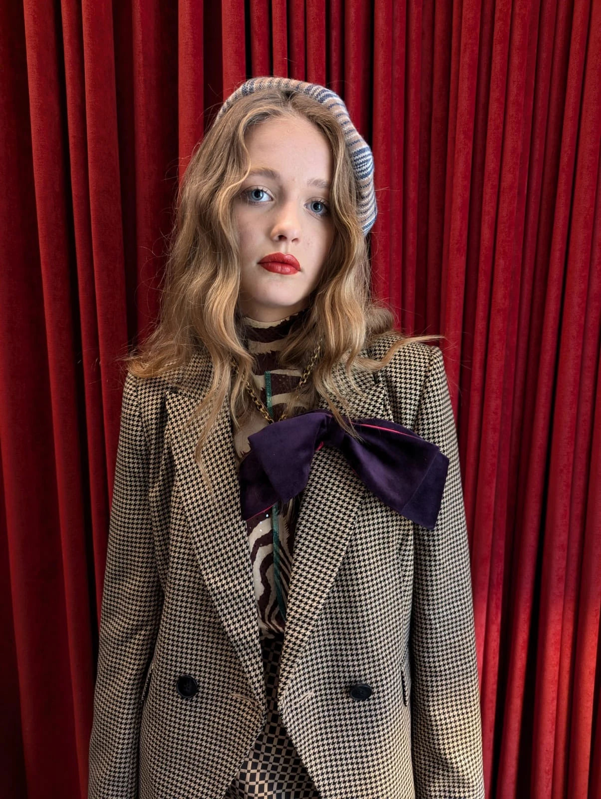 A young girl with wavy blonde hair wearing a beige and black houndstooth blazer with a large Broochella dark purple bow at the collar, a striped beret, and a patterned top, standing in front of a red velvet curtain.