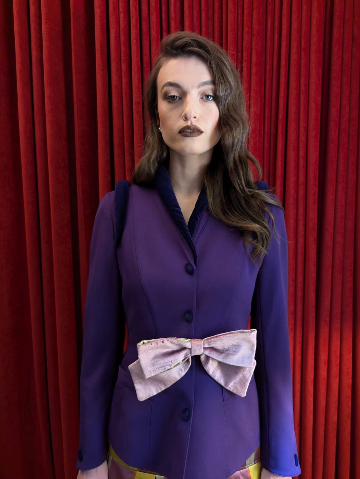 A woman stands in front of a red curtain backdrop wearing a purple tailored jacket with a large Broochella velvet pale pink bow at the waist.