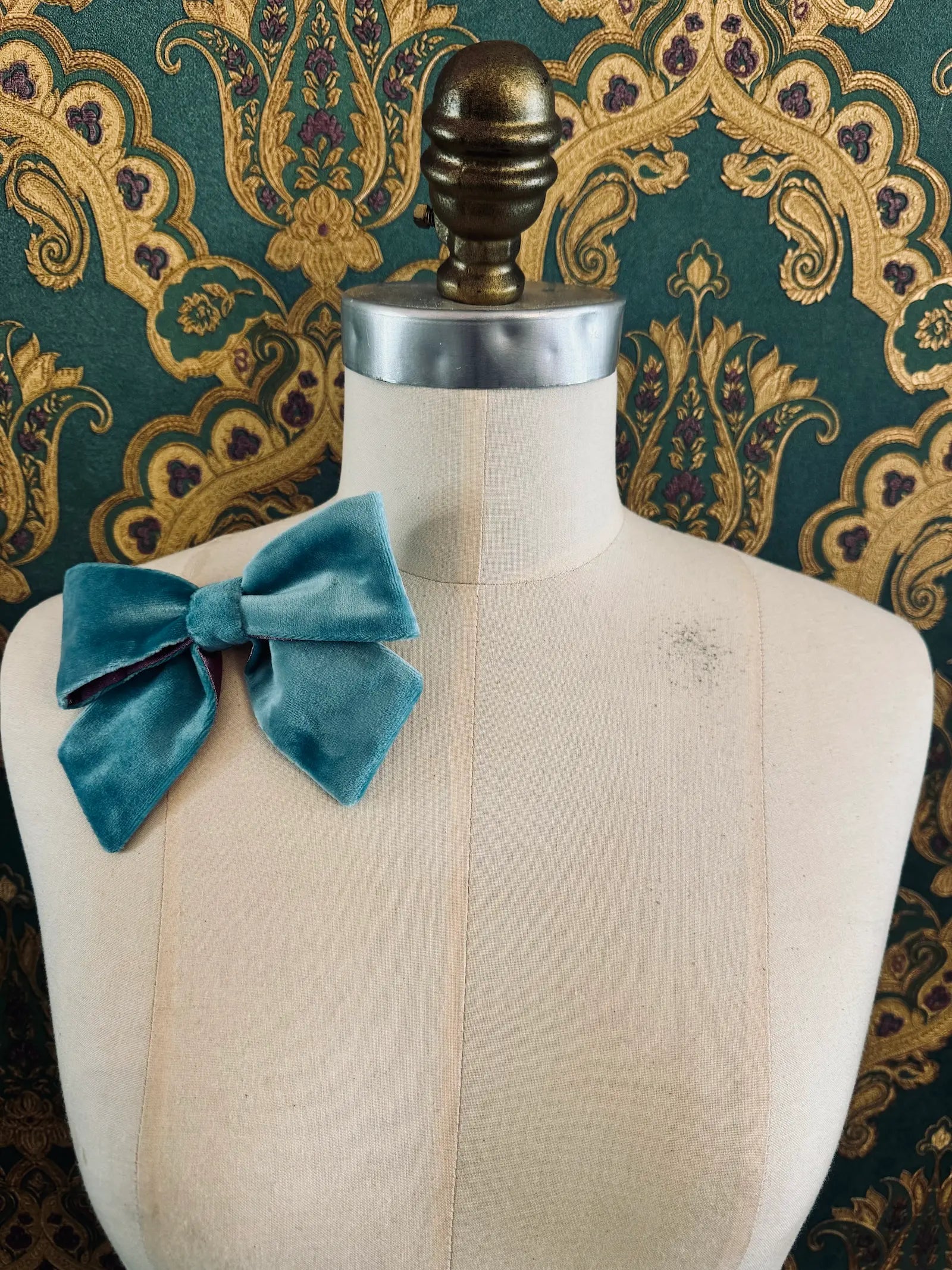 Broochella small velvet handmade bow brooch in aqua and purple. Pictured on a mannequin.  The background is green and gold damask wallpaper.