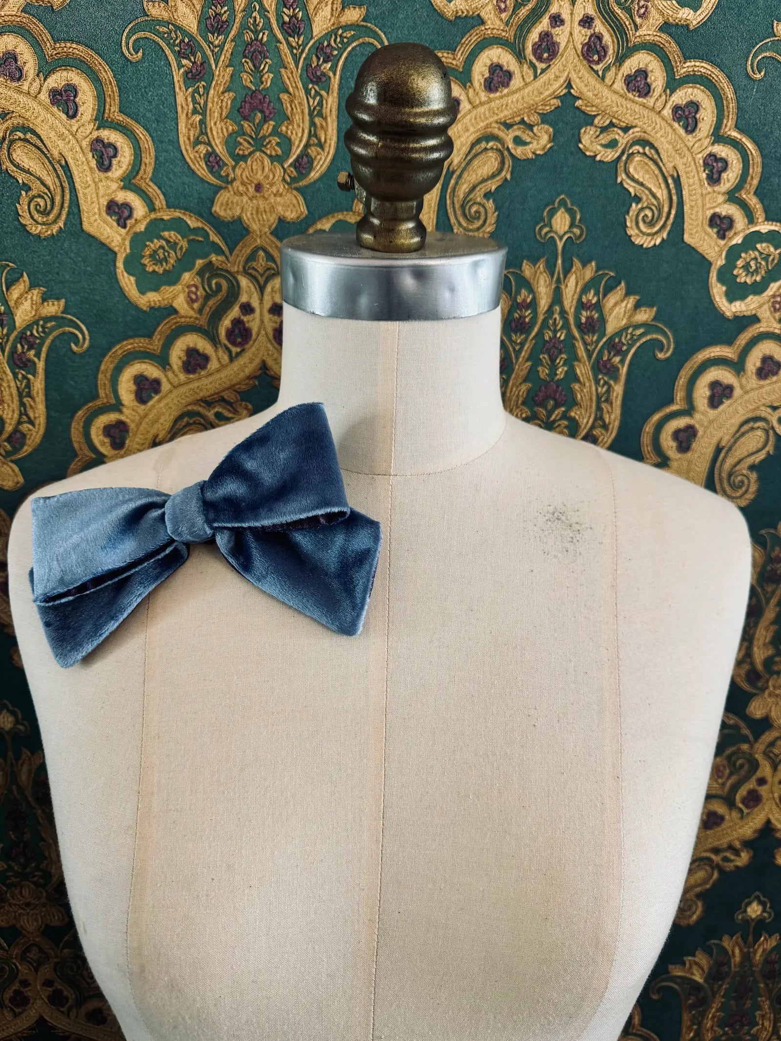 Broochella small velvet handmade bow brooch in blue and purple. Pictured on a mannequin.  The background is green and gold damask wallpaper.