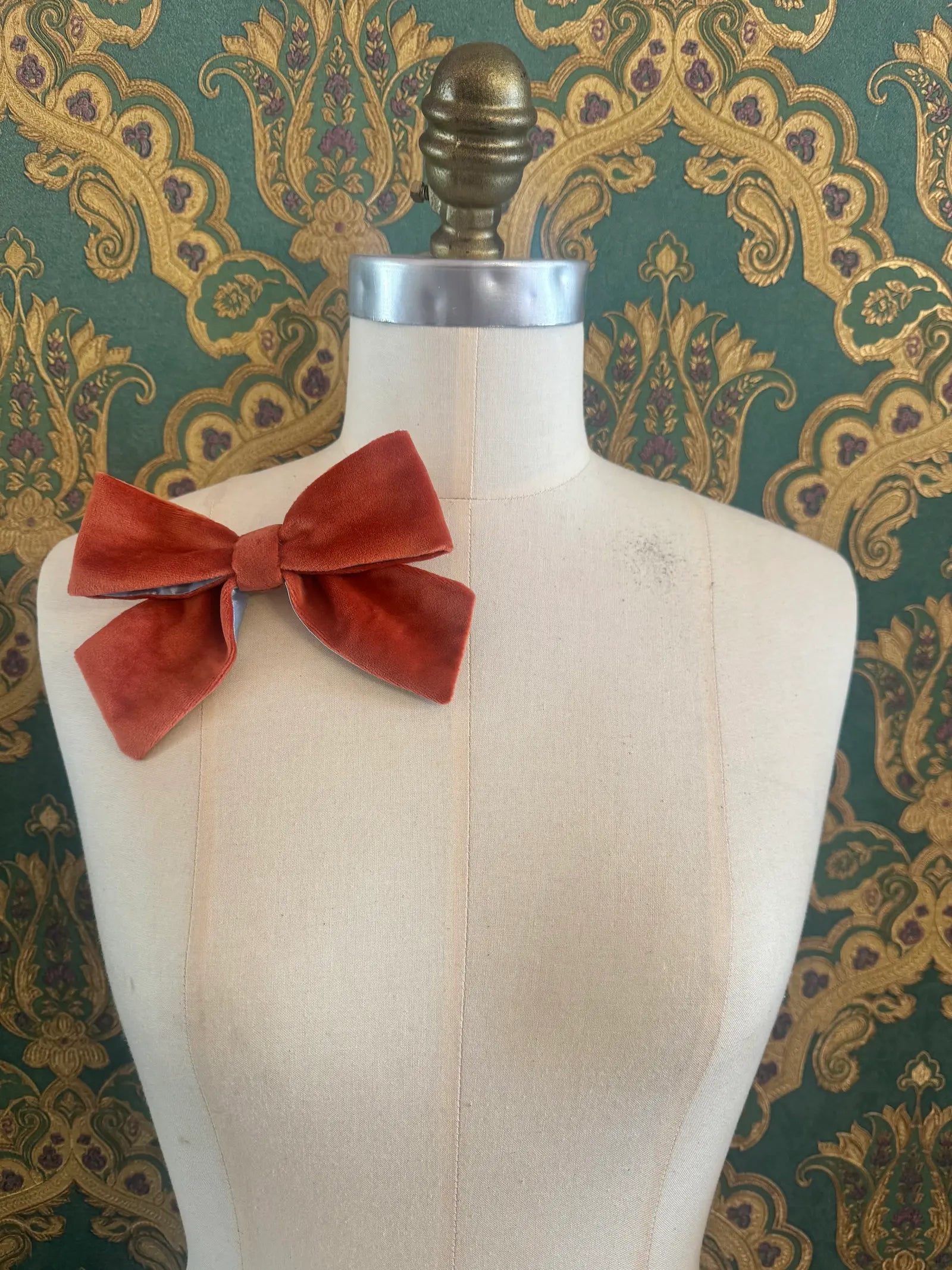 Broochella small velvet handmade bow brooch in orange and ice blue. Pictured on a mannequin.  The background is green and gold damask wallpaper.