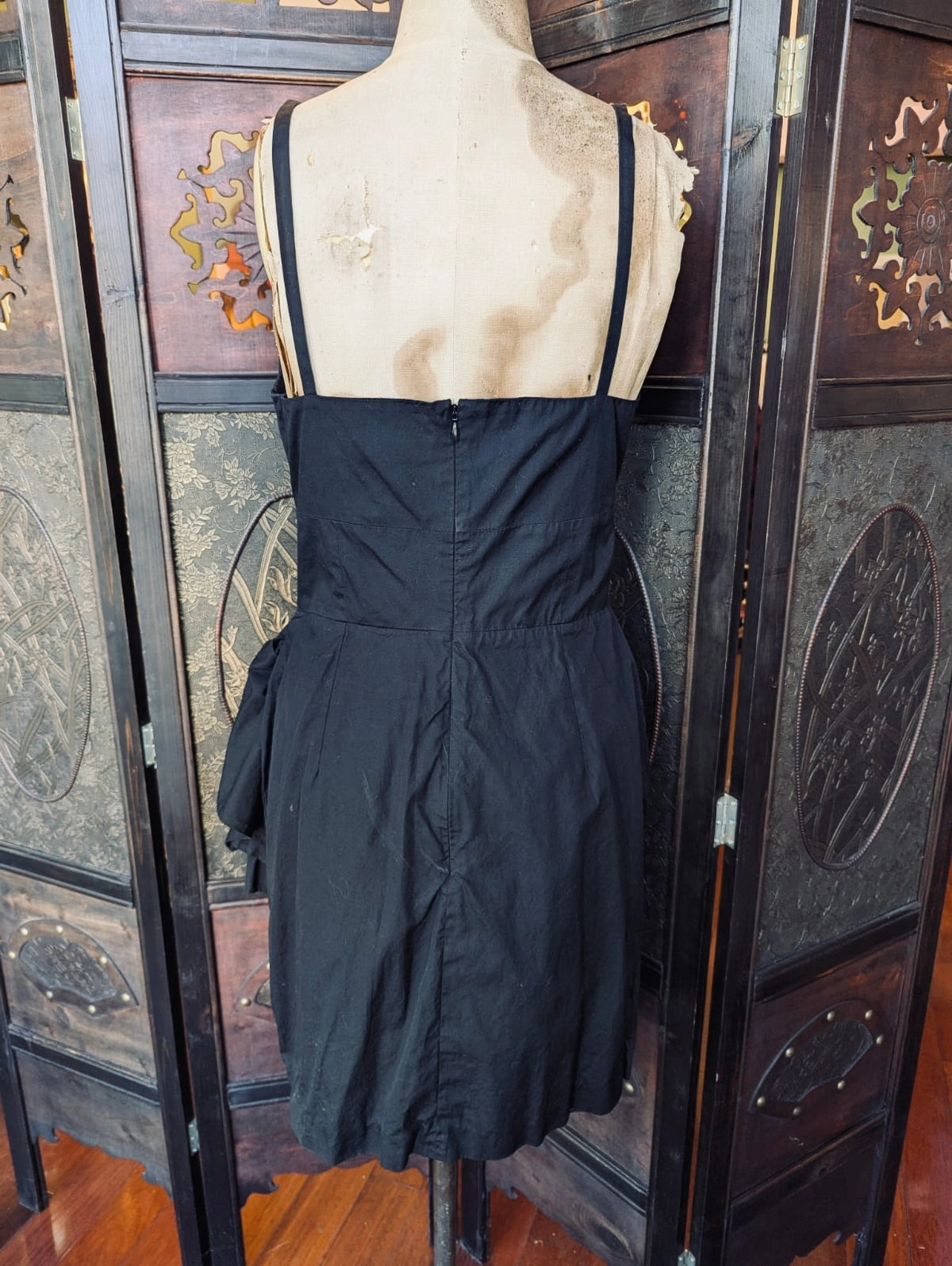 The back view of a  Broochella Victoria Beckham black sleeveless wrap-style dress with thin straps and a gathered peplum-like hem, displayed on a mannequin torso in front of a decorative wooden folding screen.