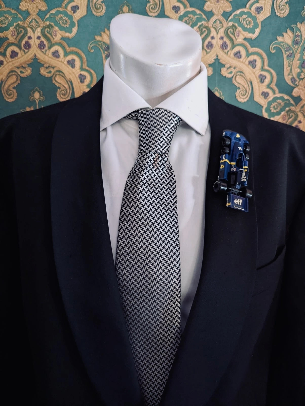 Broochella men's scuderia d'oro blu tyrrell  number 4 vintage formula one racing car brooch. Pinned on a men's mannequin wearing a black suit and a houndstooth tie. The background is green and gold damask wallpaper. 