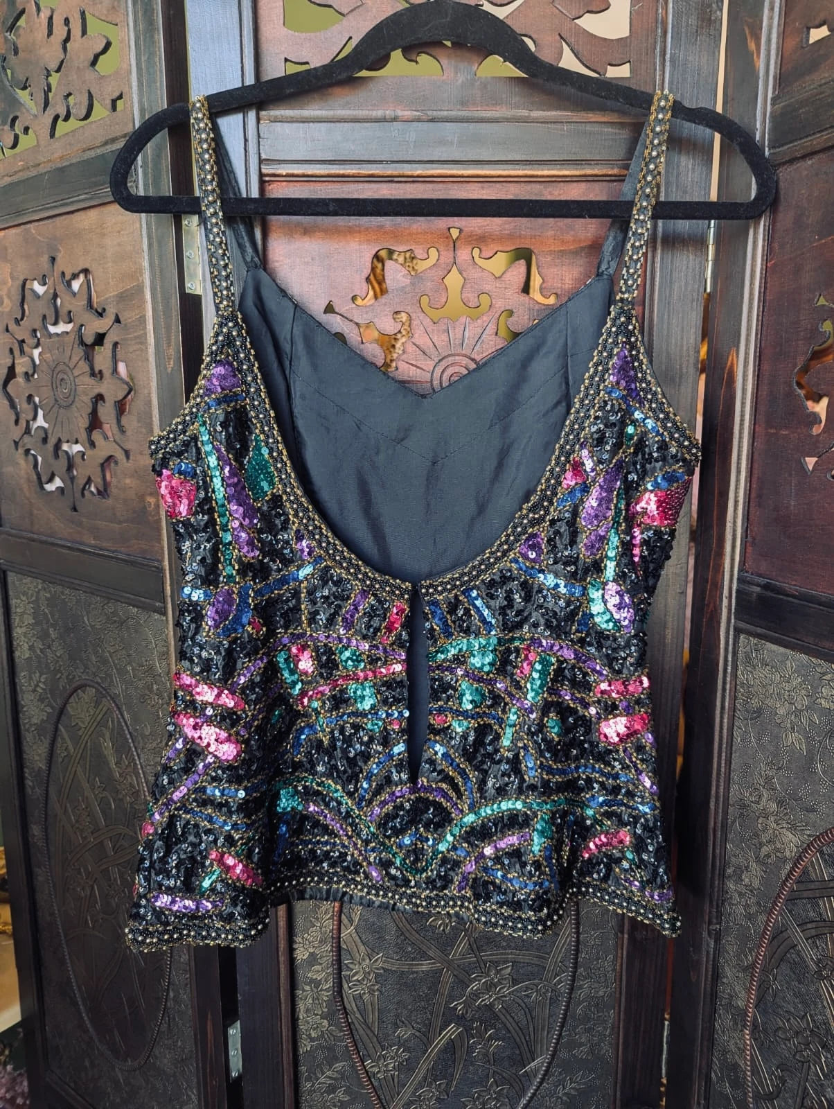 The back view of a Broochella Vintage 1980's black sequinned camisole top with multicoloured iridescent sequins and gold-tone trim, hanging on a black hanger against a dark carved wooden door.