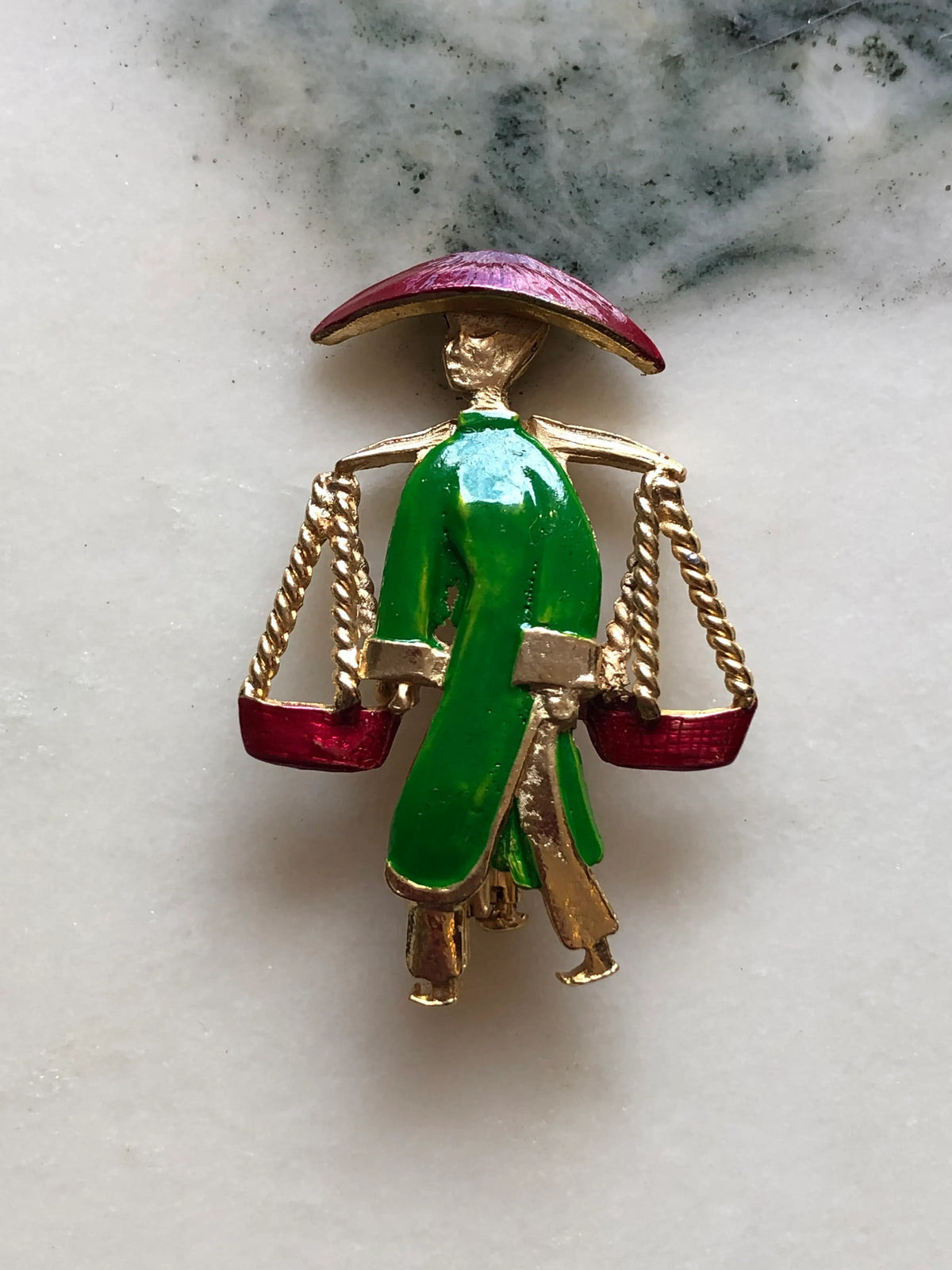 Broochella Vintage Enamel Water Carrier Brooch - Green featuring a figural water carrier in vivid green enamel with a raspberry-red hat, suspended vessels and gold-tone detailing on a cream and grey marble background.