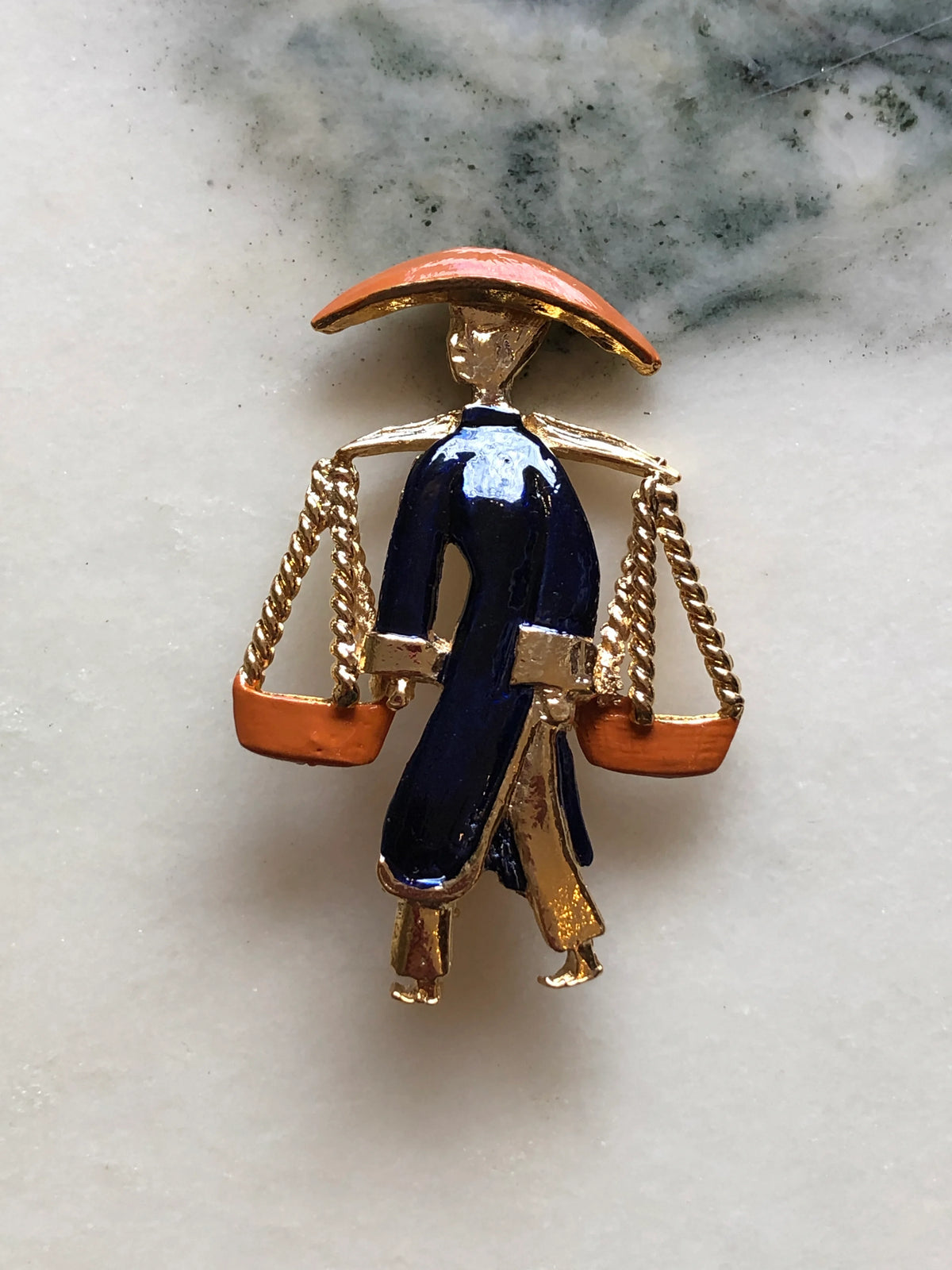 Broochella Vintage Enamel Water Carrier Brooch - Navy featuring a figural water carrier in deep navy enamel with an orange hat, suspended vessels and gold-tone detailing on a cream and grey marble background.