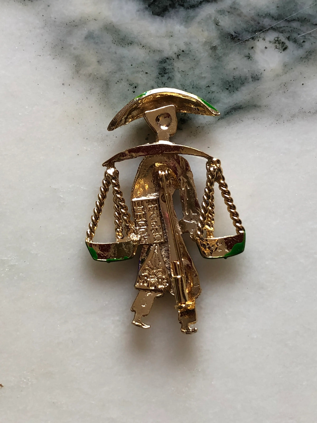 The back view of Broochella Vintage Enamel Water Carrier Brooch - Purple featuring a figural water carrier in violet-purple enamel with a green hat, suspended green vessels and gold-tone detailing on a cream and grey marble background.