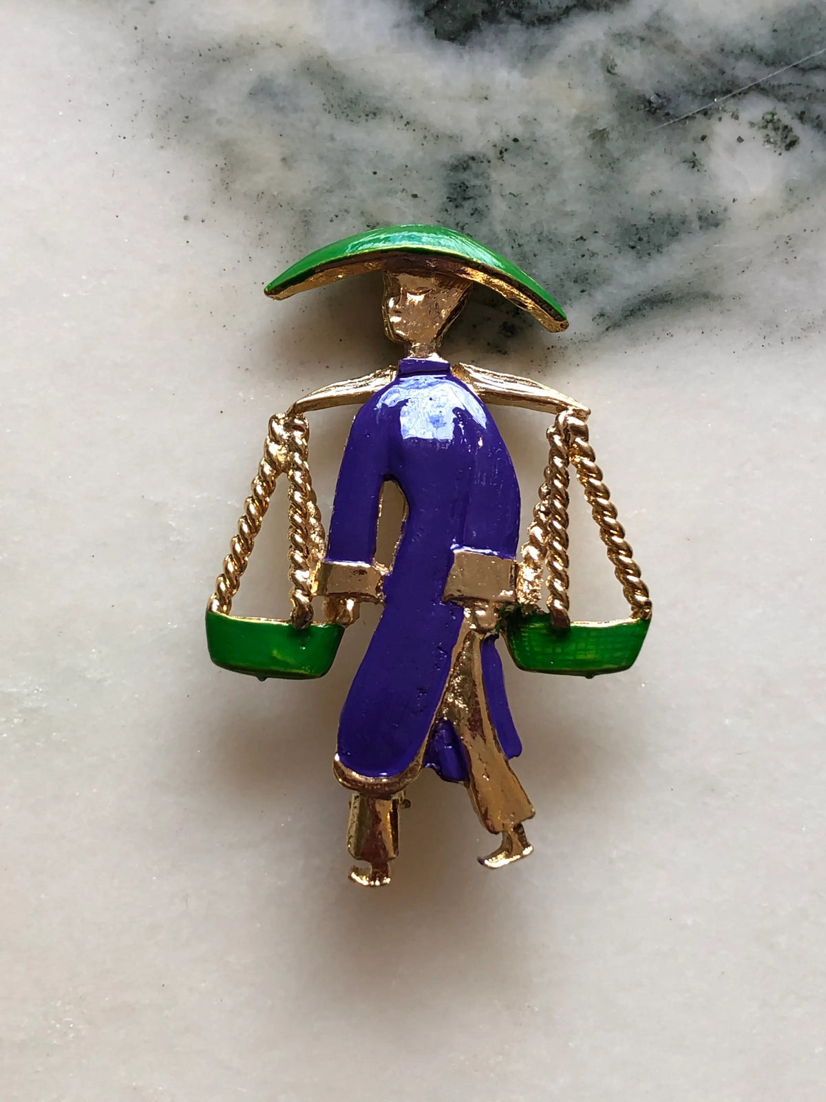 Broochella Vintage Enamel Water Carrier Brooch - Purple featuring a figural water carrier in violet-purple enamel with a green hat, suspended green vessels and gold-tone detailing on a cream and grey marble background.