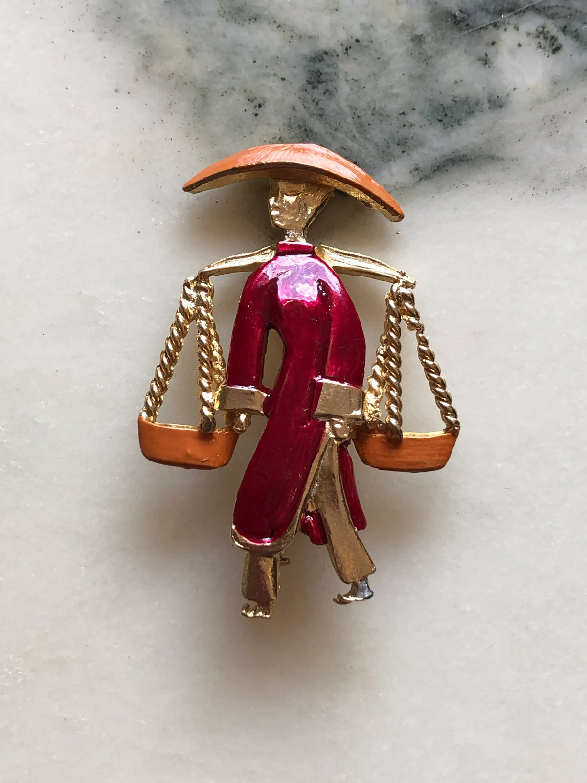 Broochella Vintage Enamel Water Carrier Brooch featuring a figural water carrier in raspberry-red and orange enamel with suspended vessels and gold-tone detailing on a cream and grey marble background.