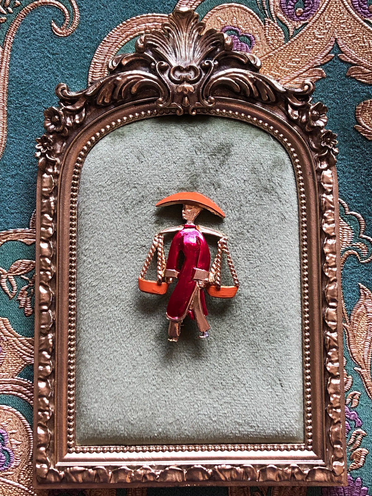 Broochella Vintage Enamel Water Carrier Brooch featuring a figural water carrier in raspberry-red and orange enamel with suspended vessels and gold-tone detailing. Pictured on a green velvet tray with an ornate gold frame. The background is green and gold damask wallpaper