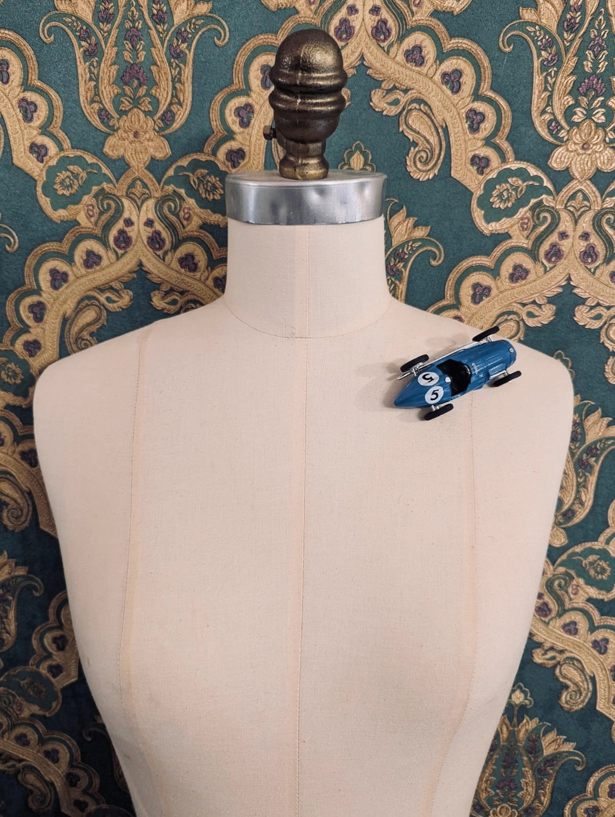 Broochella  scuderia d'oro blu veloce number five blue vintage formula one racing car brooch. Pinned on a  mannequin. The background is green and gold damask wallpaper. 