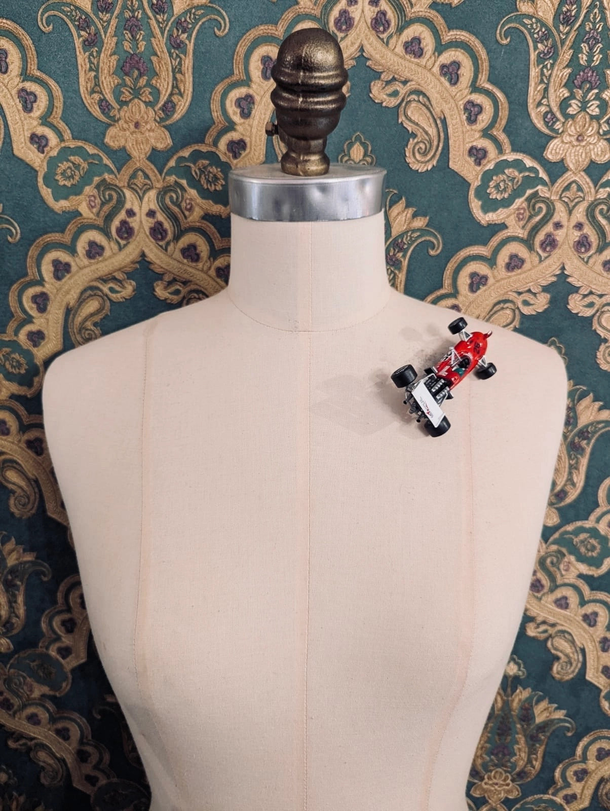 Broochella  scuderia d'oro rosso turbo 26 red, vintage formula one racing car brooch. Pinned on a mannequin. The background is green and gold damask wallpaper. 