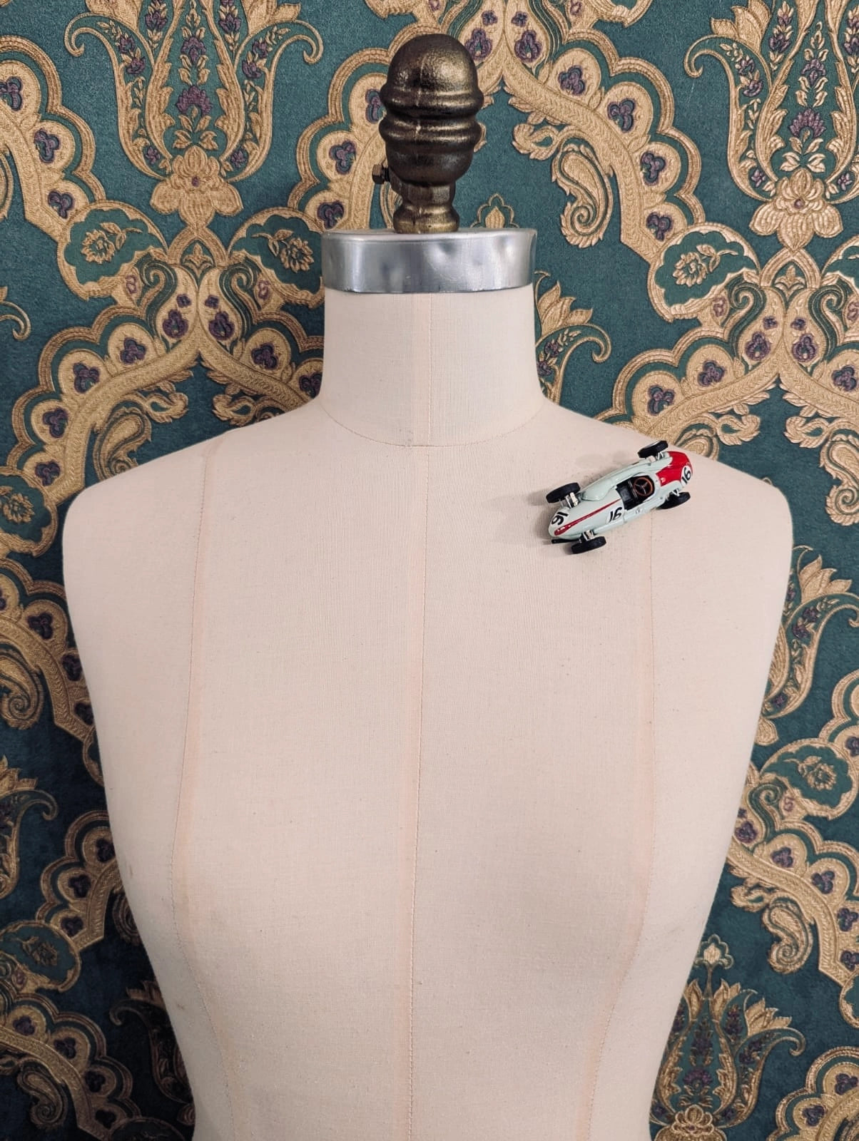 Broochella  scuderia d'oro verde corsa number 16 green, vintage formula one racing car brooch. Pinned on a mannequin. The background is green and gold damask wallpaper. 