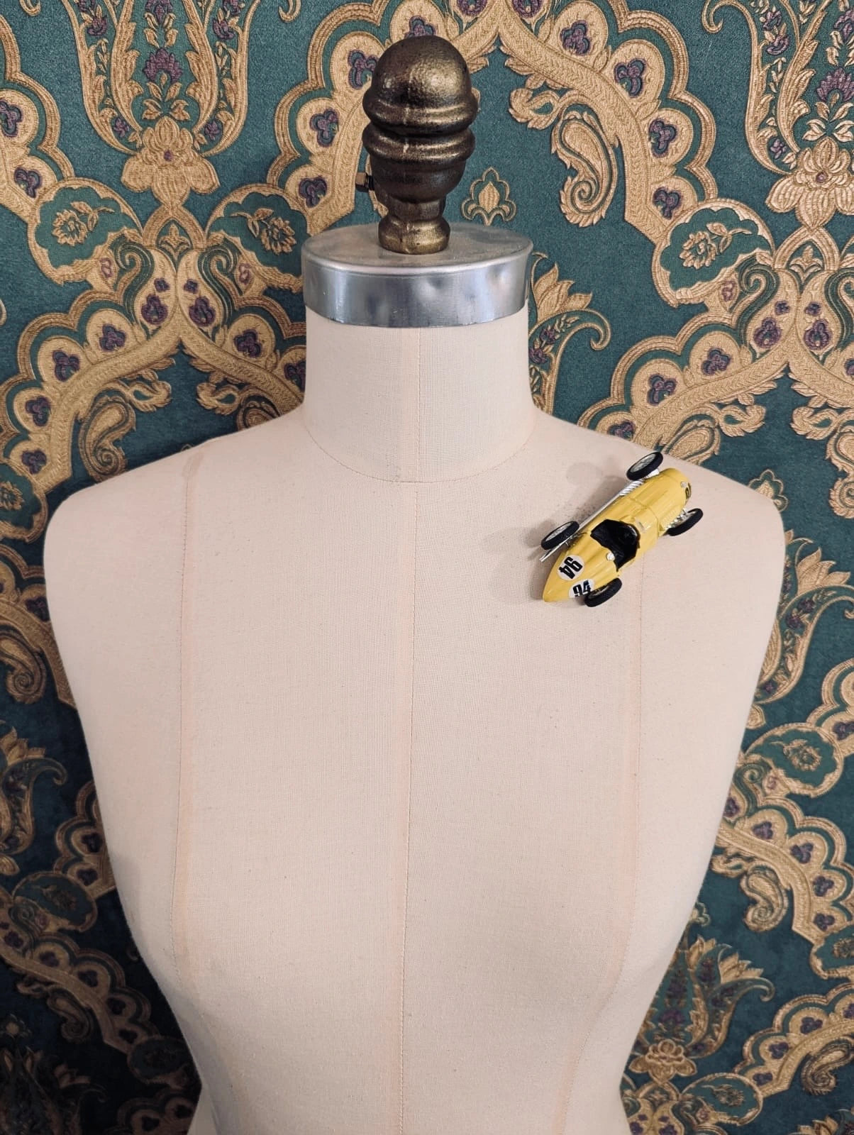 Broochella  scuderia d'oro giallo fuoco 94 yellow, vintage formula one racing car brooch. Pinned on a mannequin. The background is green and gold damask wallpaper. 