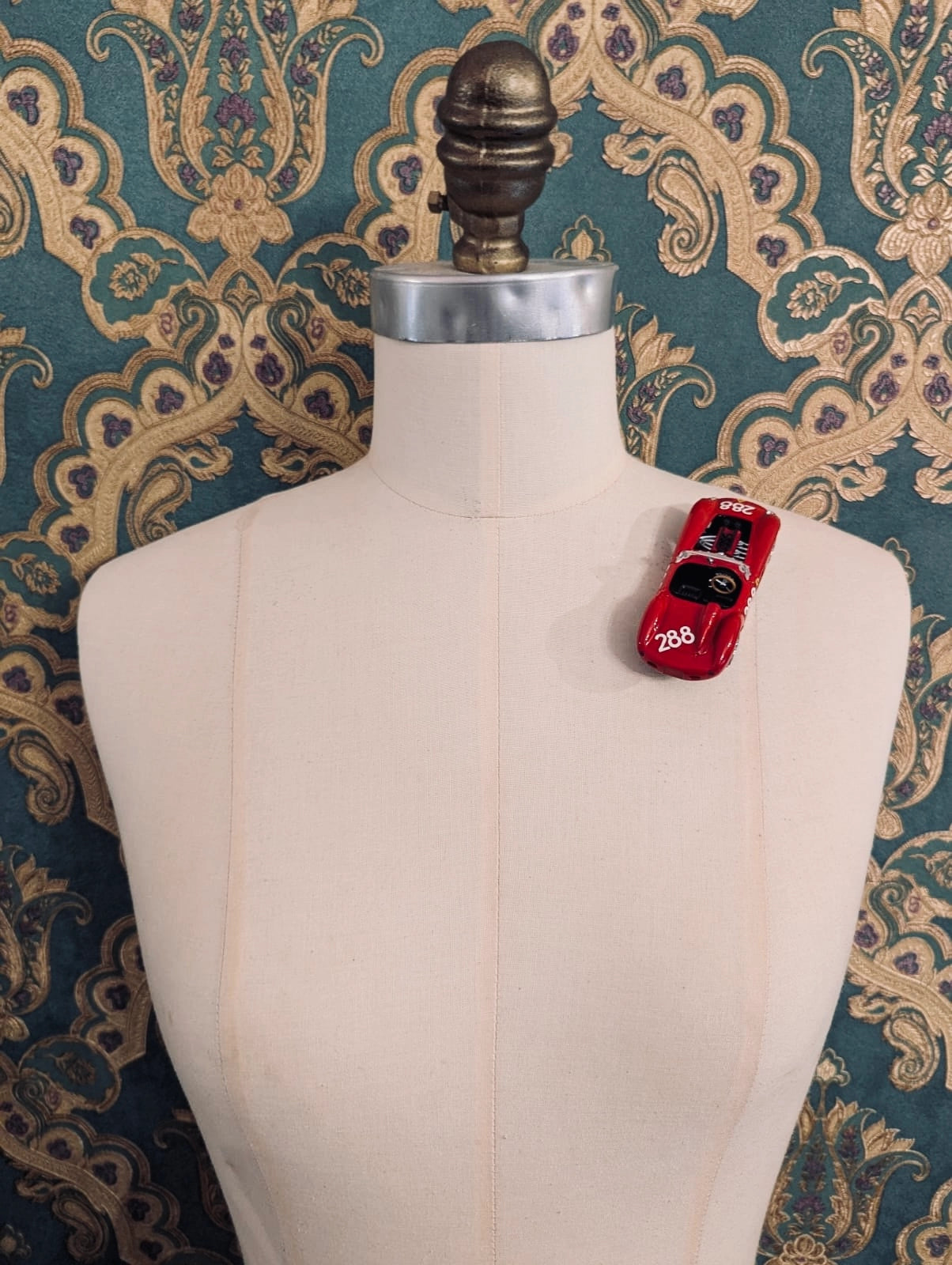 Mannequin torso with a Broochella rosso corsa number 288  vintage formula one brooch  car on a mannequin. The background green and gold damask wallpaper. 