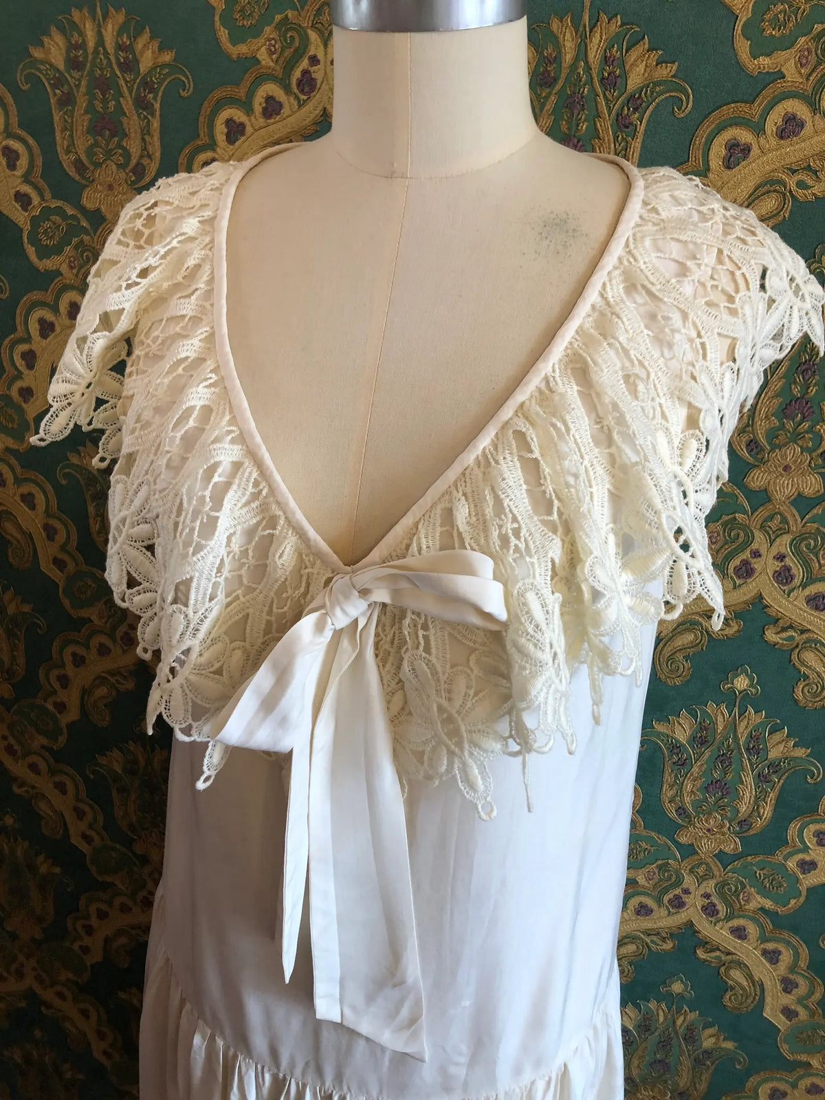Broochella vintage Ninette ivory silk maxi dress with an ornate lace cape collar, deep V-neckline and satin bow, labelled vintage AU 12 and approximately modern AU 8. Pictured on a mannequin against green and gold damask wallpaper.