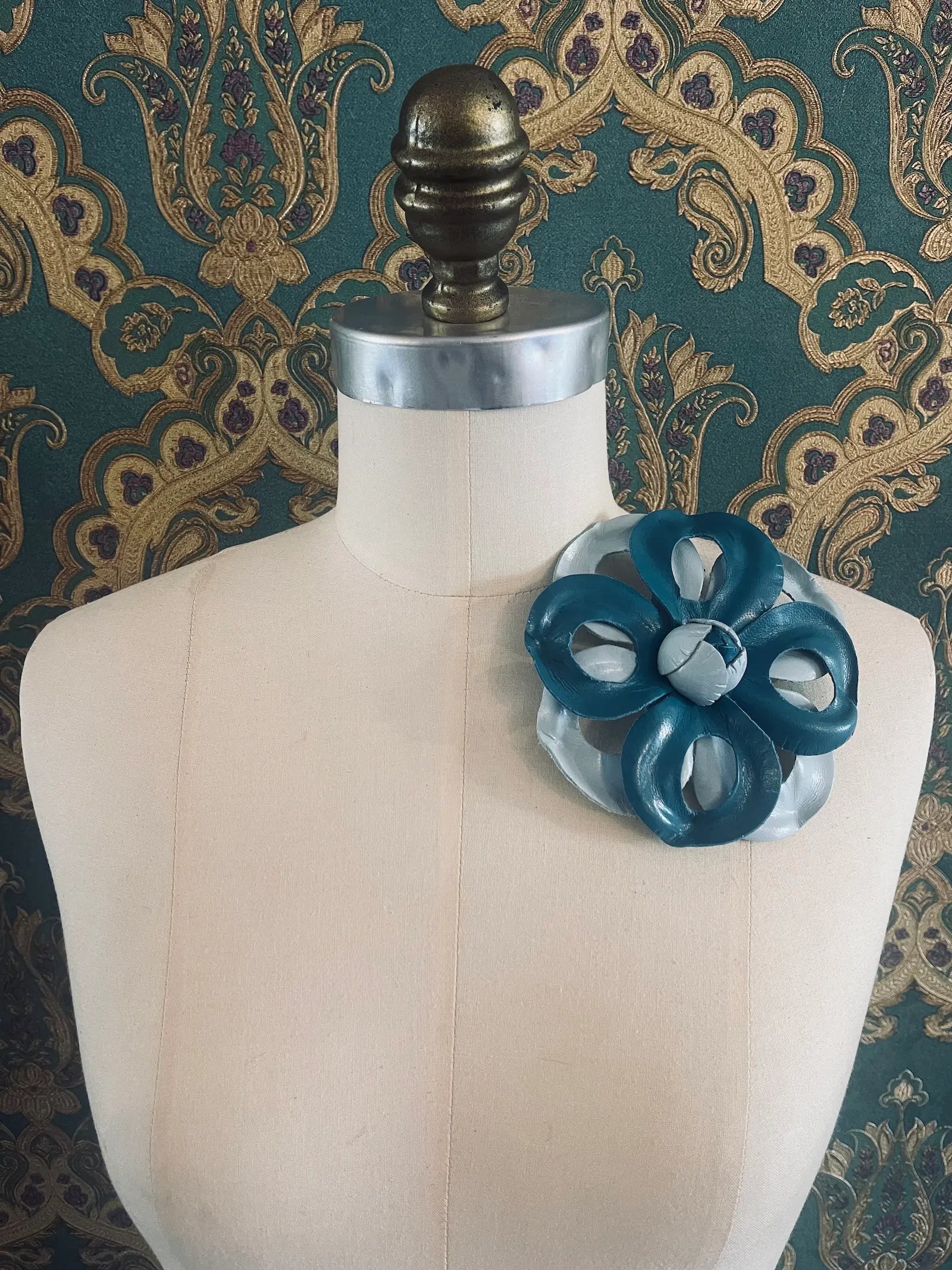 Broochella viola del pensiero handmade two tone blue leather flower brooch. Styled on a mannequin. The background is green and gold damask wallpaper