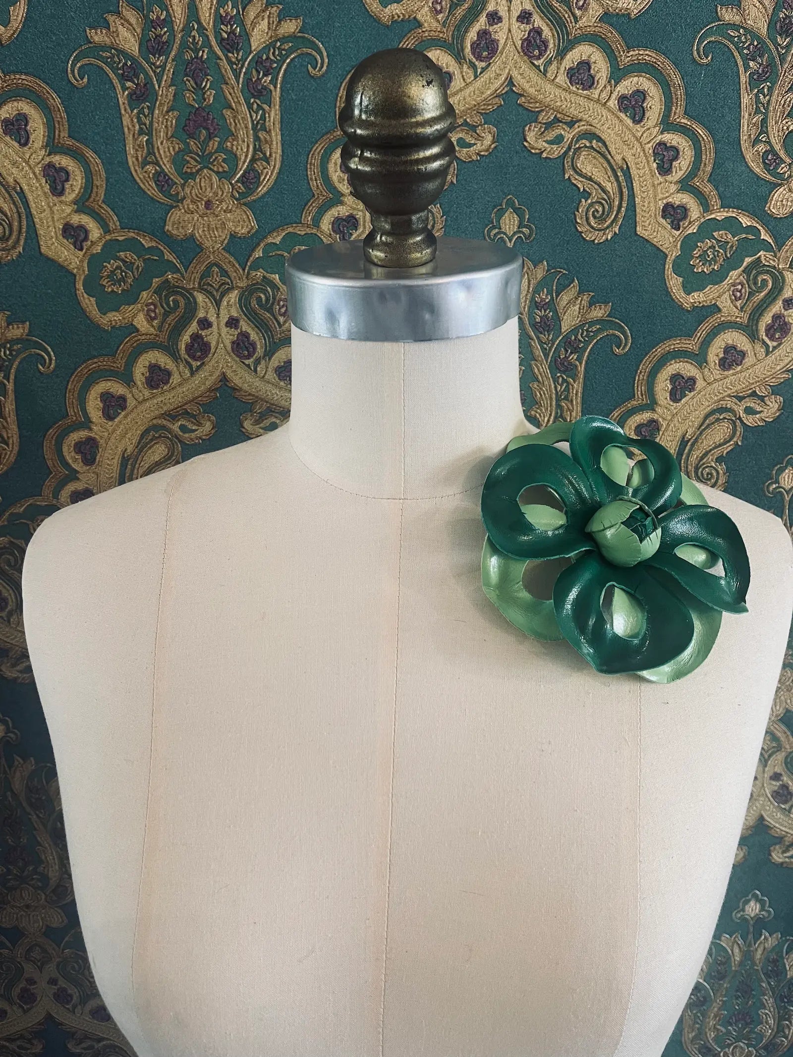 Broochella viola del pensiero handmade two tone green leather flower brooch. Styled on a mannequin. The background is green and gold damask wallpaper