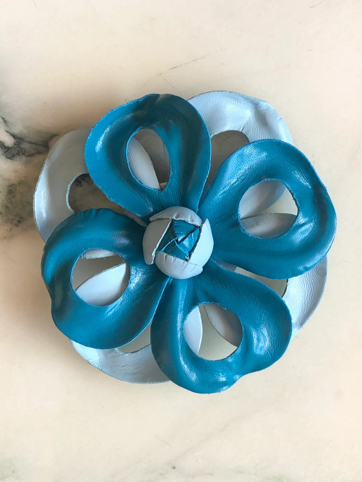 Broochella Men's Viola del Pensiero Leather Flower Brooch Pin in blue, featuring sculptural two-tone turquoise and pale blue leather petals with open cut-out detailing and a handcrafted centre on a cream marble background.