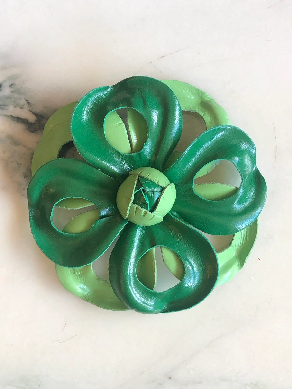 Broochella Men's Viola del Pensiero Leather Flower Brooch Pin in green, featuring sculptural two-tone dark and light green leather petals with open cut-out detailing and a handcrafted centre on a cream marble background.