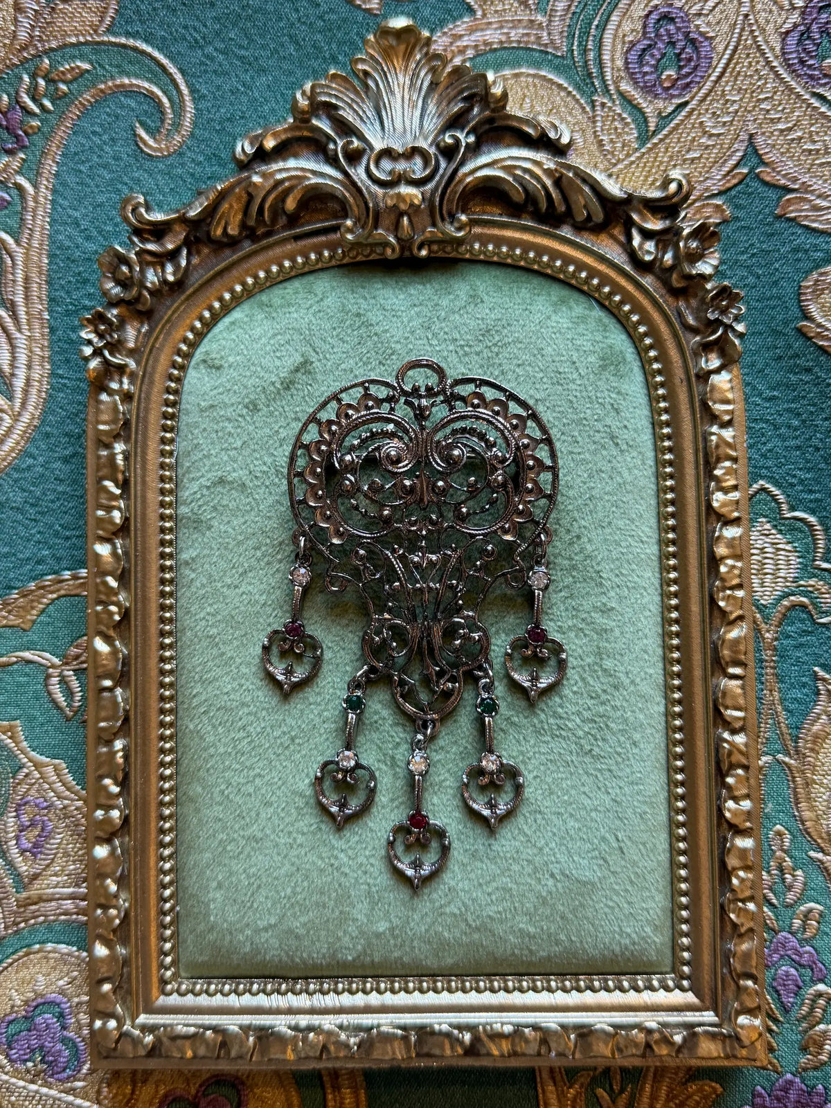 Broochella Vittoria Vintage Silver Filigree Drop Brooch featuring ornate silver-tone openwork, five articulated drops and green, red and clear stones. Pictured on a green velvet tray with an ornate gold frame. The background is green and gold damask wallpaper.