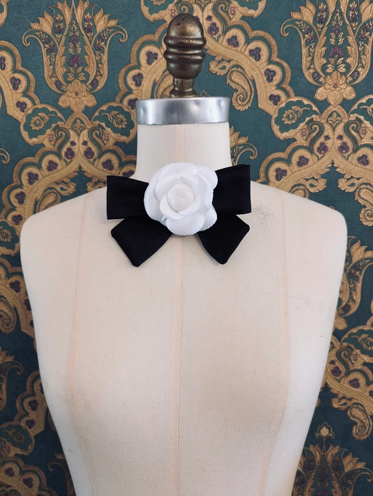 Broochella viva l'amor handmade silk black bow brooch with white flower centre.  It is pinned to a mannequin. The background is green and gold damask wallpaper.