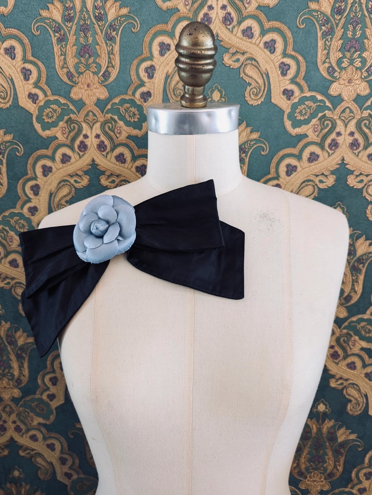 Broochella viva l'amor handmade black silk bow brooch, with white flower embellishment.  The background is green and gold damask wallpaper.
