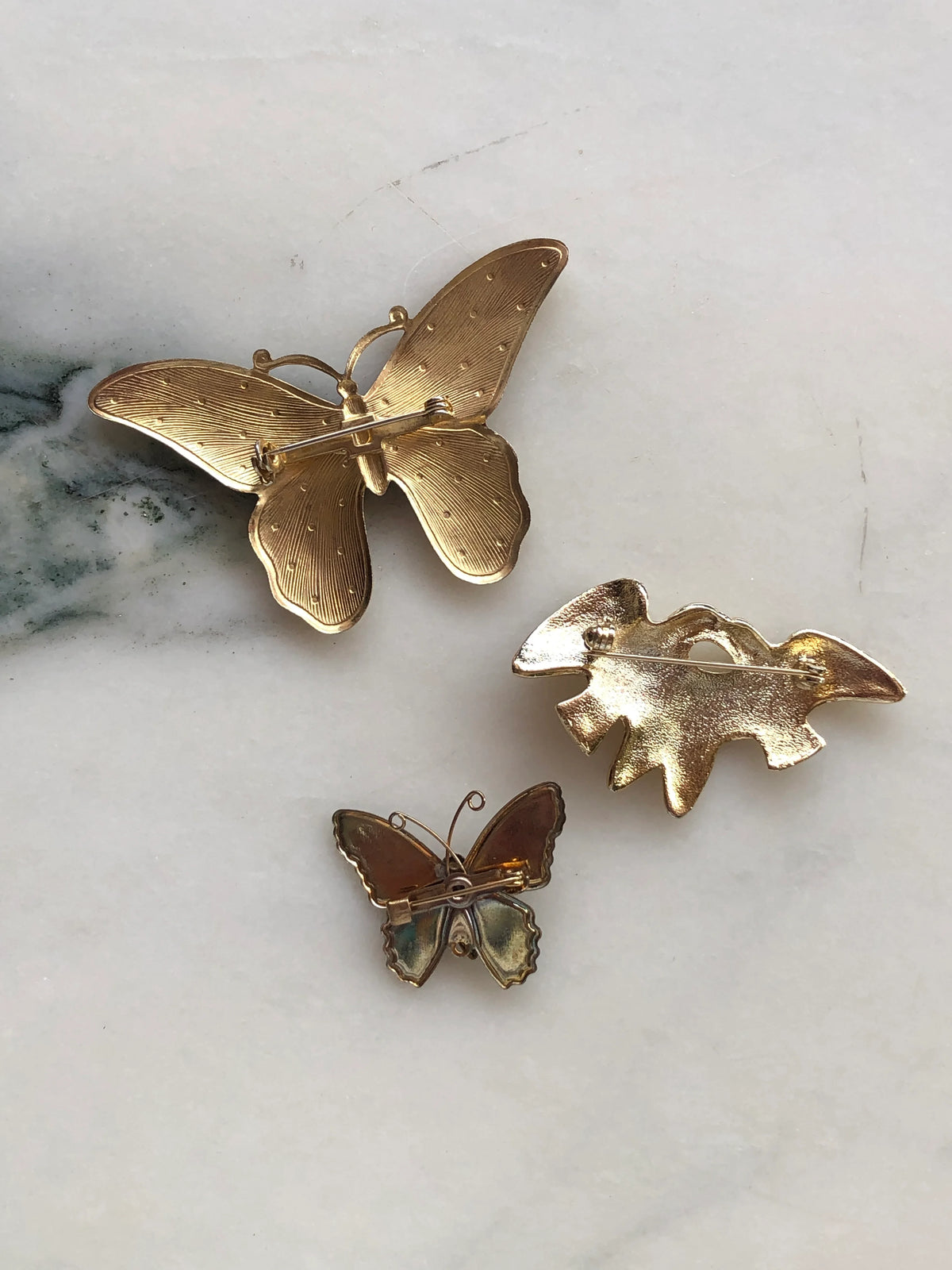 Broochella Wings of Change Vintage Brooch Set shown from the back, displaying the gold-tone metal finishes, pin fastenings and clasps of the two butterfly brooches and bird brooch, photographed on a pale white marble background with grey veining.