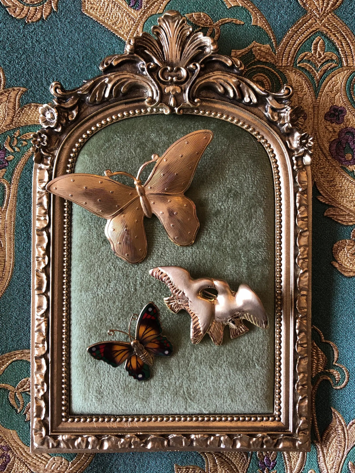 Wings of Change Vintage Brooch Set featuring two butterfly brooches and a gold bird brooch displayed in an ornate vintage gold frame with green velvet, against a teal and gold embroidered fabric background.