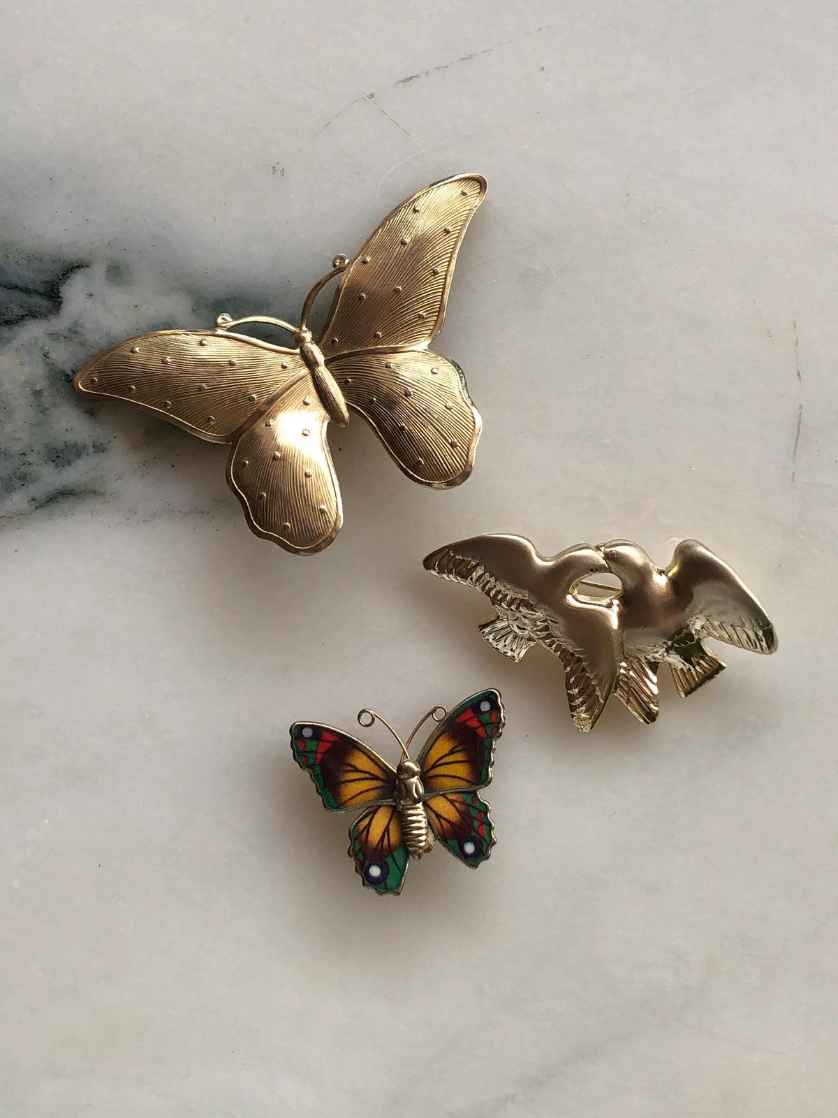 Broochella Wings of Change Vintage Brooch Set featuring a large gold butterfly, colourful enamel butterfly and sculptural gold bird brooch, photographed on a pale white marble background with grey veining.