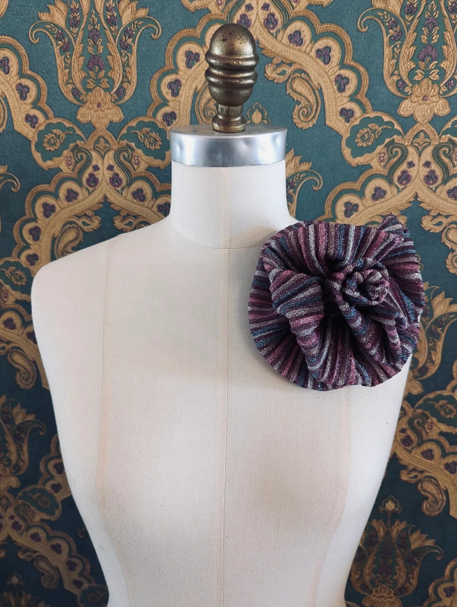Broochella wonka handmade flower brooch in pink and purple striped lurex. It is pinned to a mannequin. The background is green and gold damask wallpaper.
