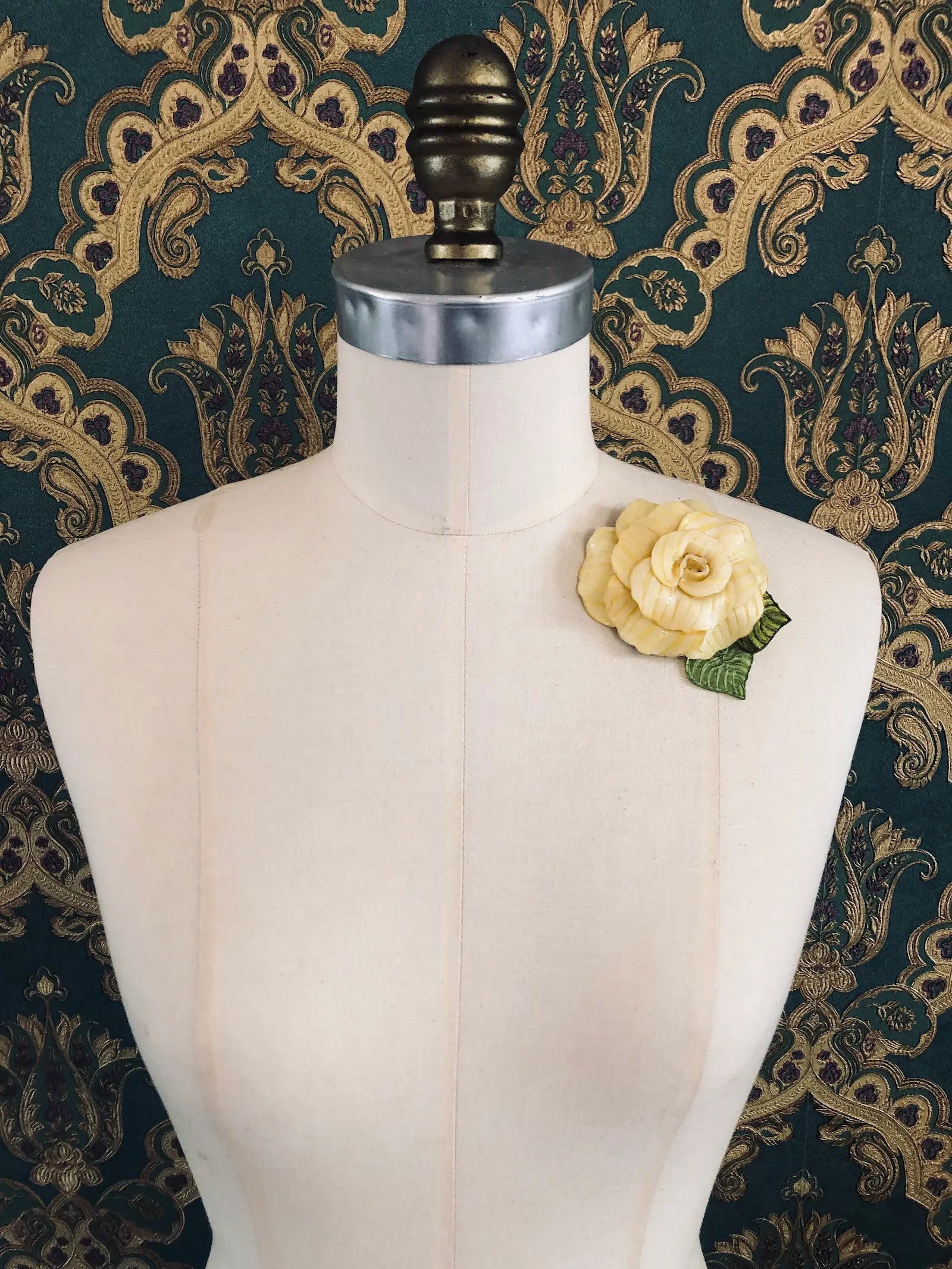 A Broochella x Fabrice Paris yellow moulded plastic rose brooch with green leaves, pinned to the chest of a cream-coloured dress form against a dark green and gold patterned damask backdrop.