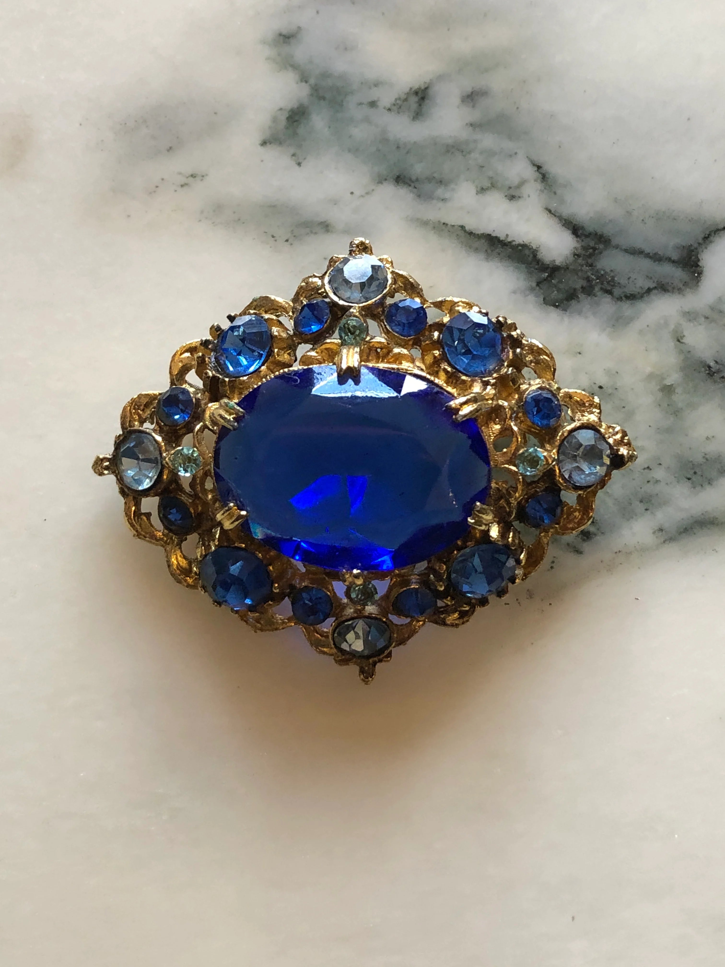 Broochella Zaffiro vintage gold-tone brooch with a large cobalt-blue centre crystal and blue rhinestone surround. Pictures on a white marble surface