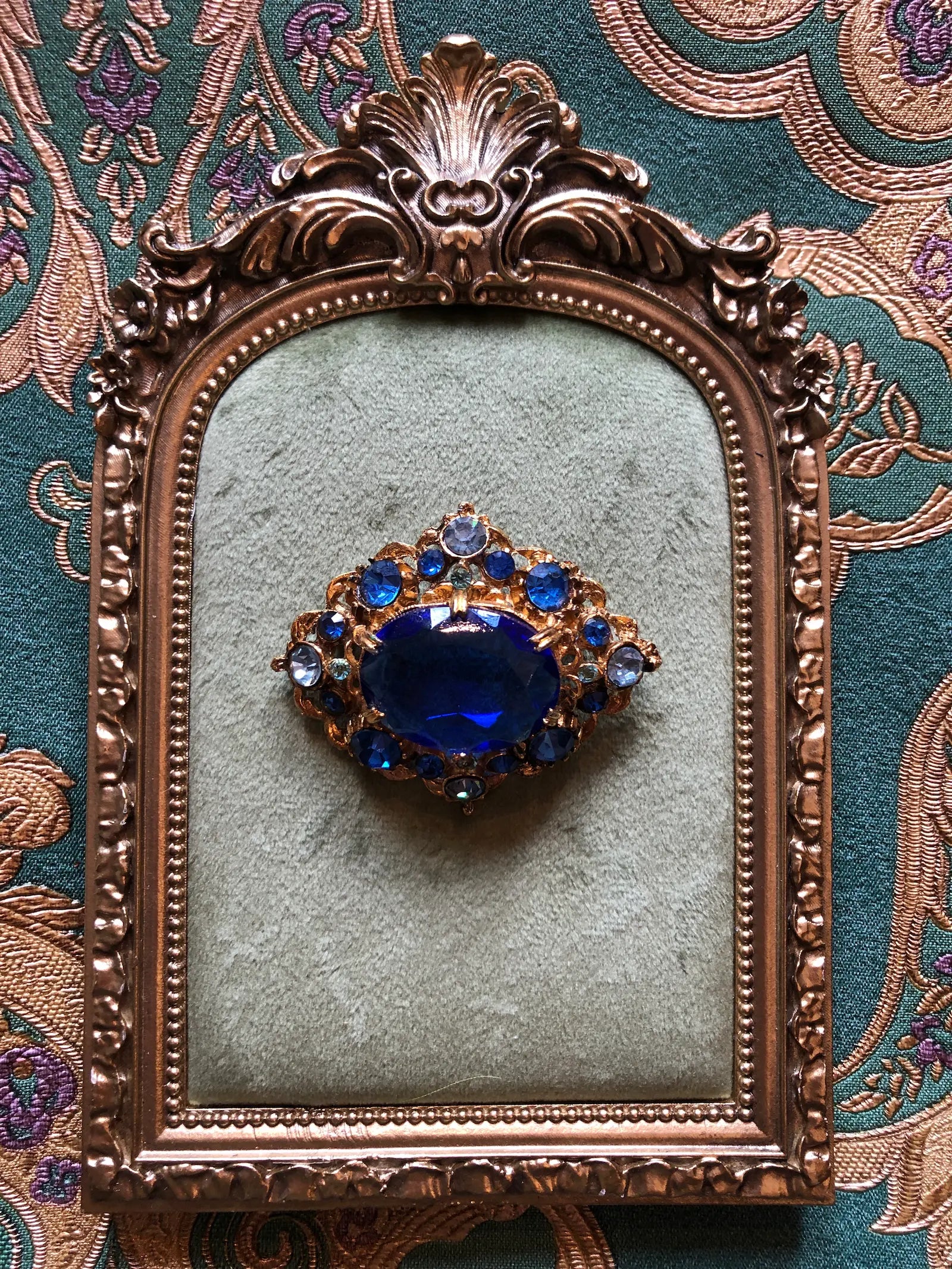 A Broochella Zaffrio vintage brooch featuring a large oval deep blue sapphire surrounded by smaller round blue and clear crystals in a gold-toned setting, displayed on a light green velvet cushion inside an antique bronze mirror frame.