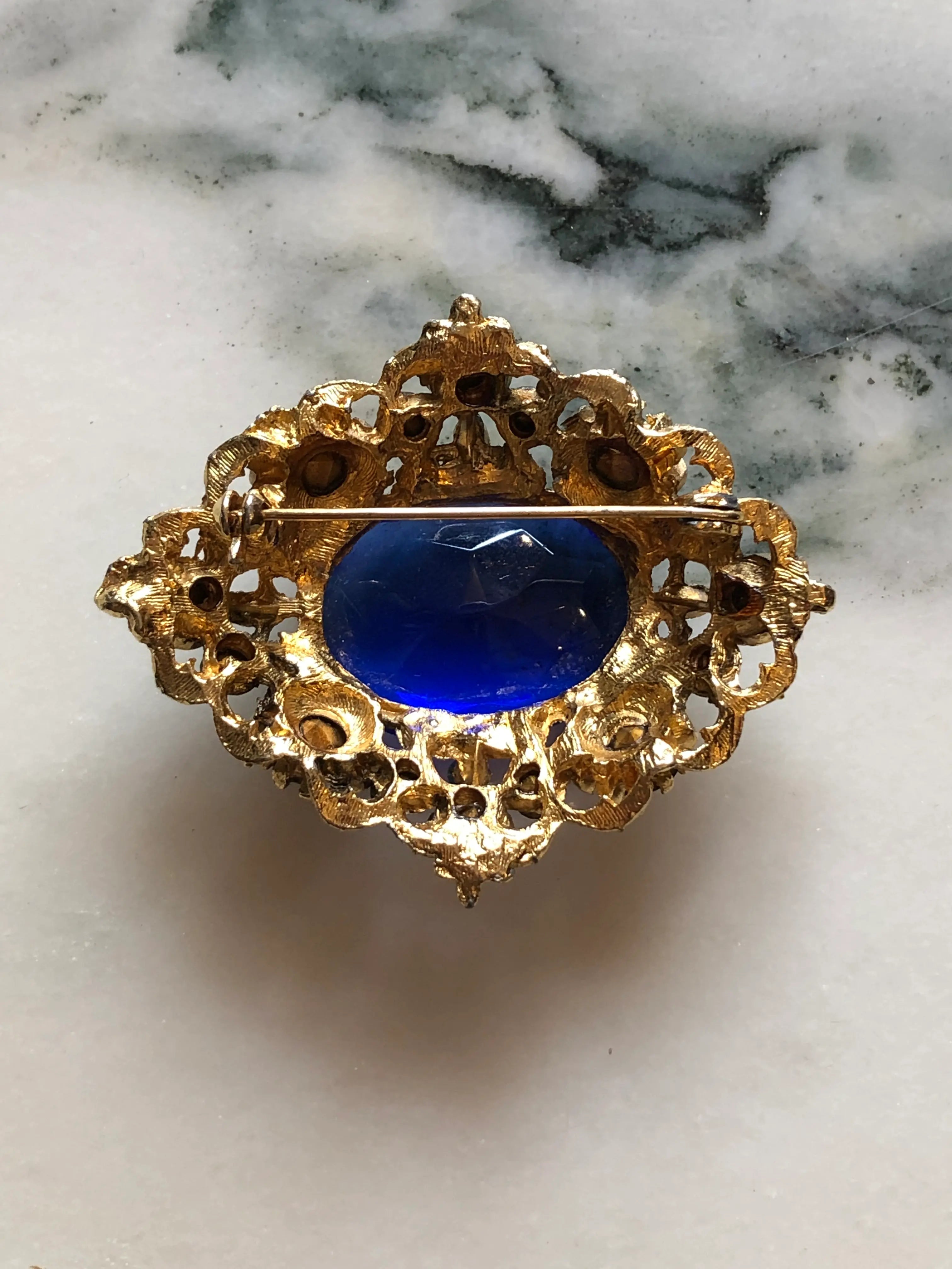 The back view of Broochella Zaffiro vintage gold-tone brooch with a large cobalt-blue centre crystal and blue rhinestone surround. Pictures on a white marble surface