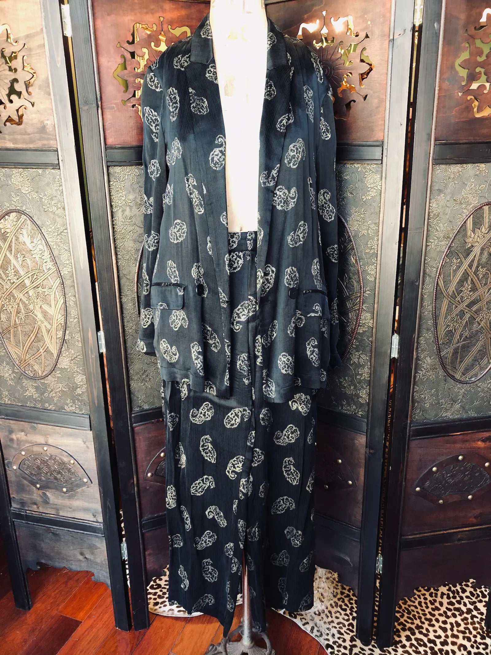 Broochella Zara, Small, Two-piece suit set
Matching blazer and trousers. It is black with paisley print and a Silky crinkle-texture fabric. It is presented on a vintage mannequin The background is an ornate, dark wood room divider. 