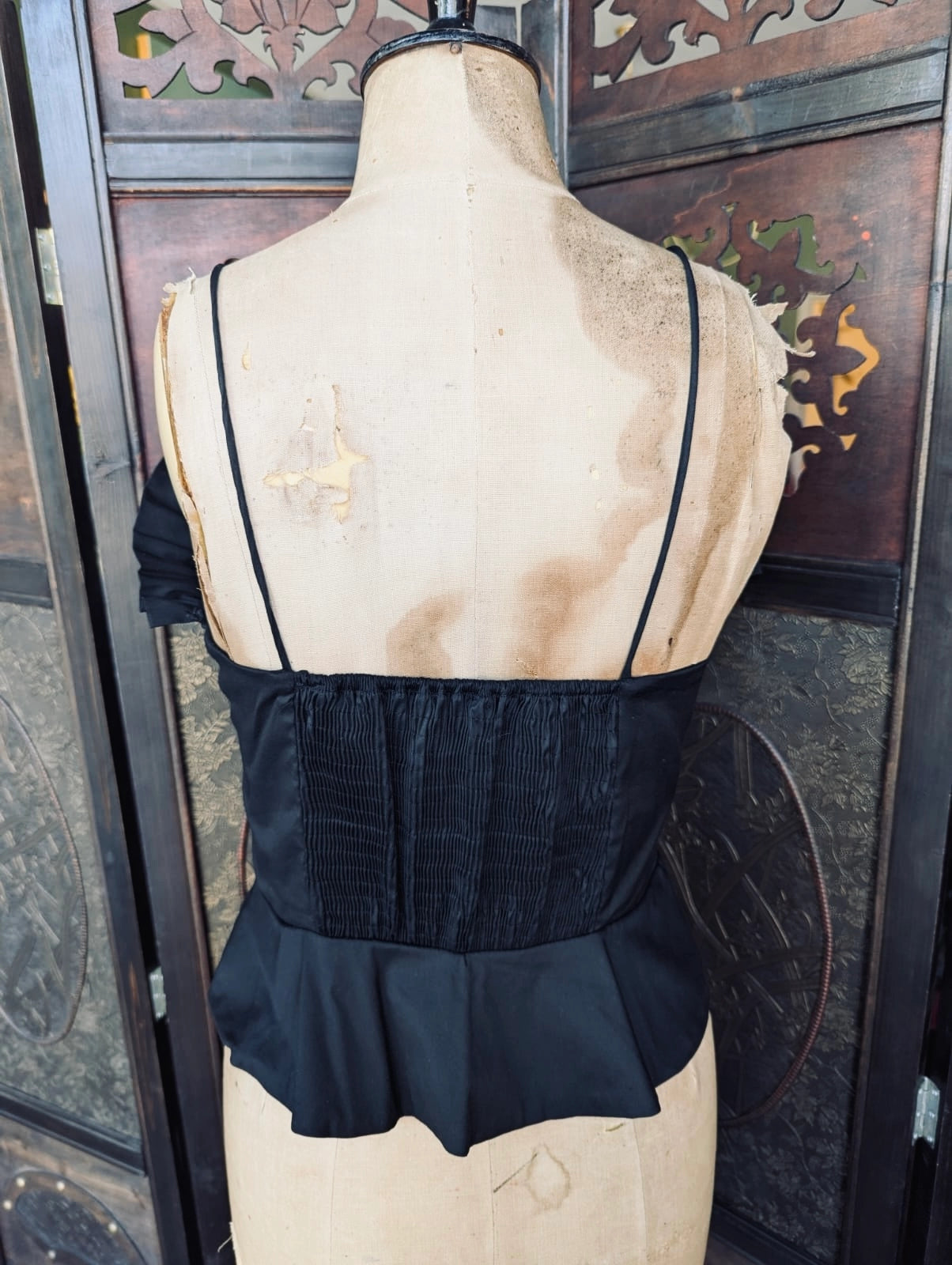 The back view of a Broochella Zara black ruffle peplum cami top in black. Pictured on a mannequin. The background is an ornate dark wood room divider