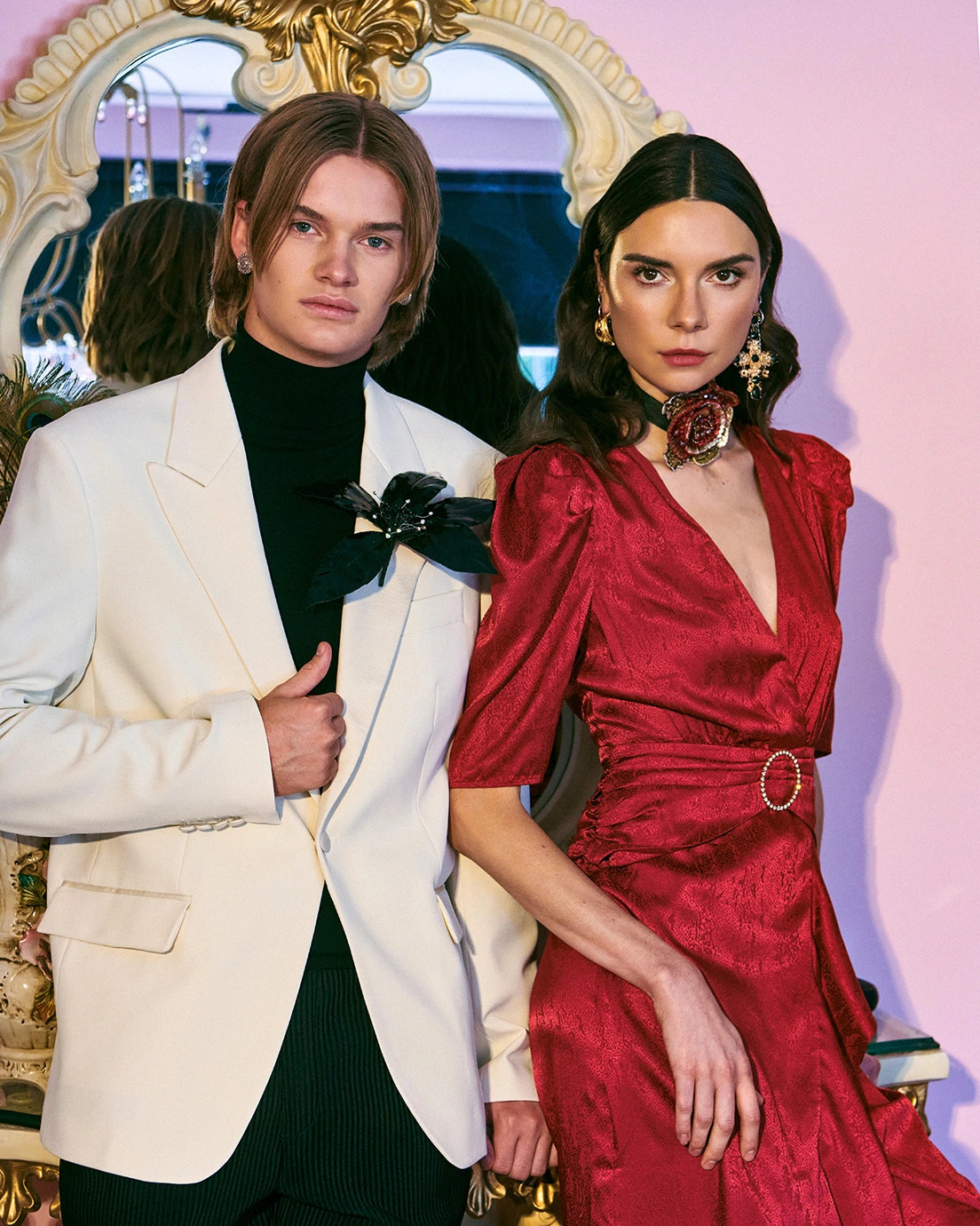 A man and a woman pose together in an elegant indoor setting. The man wears a cream-coloured blazer over a black turtleneck, and the woman wears a deep red satin dress with a V-neckline, puffed short sleeves, and a decorative belt. He is wearing the Broochella Giglio, black patent leather flower brooch. 