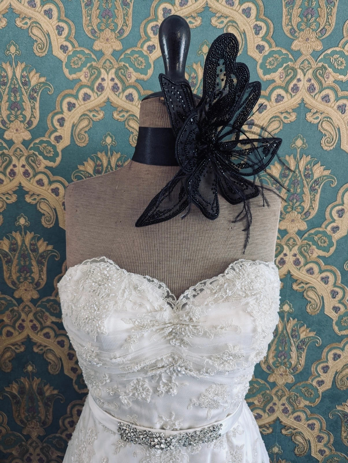 Broochella azalea large bridal, hand beaded, black, large flower brooch, with lace and crystal brooch embellishment. Pinned on a mannequin. The mannequin is dressed in a white, beaded, strapless wedding dress. The background is green and gold damask wallpaper.