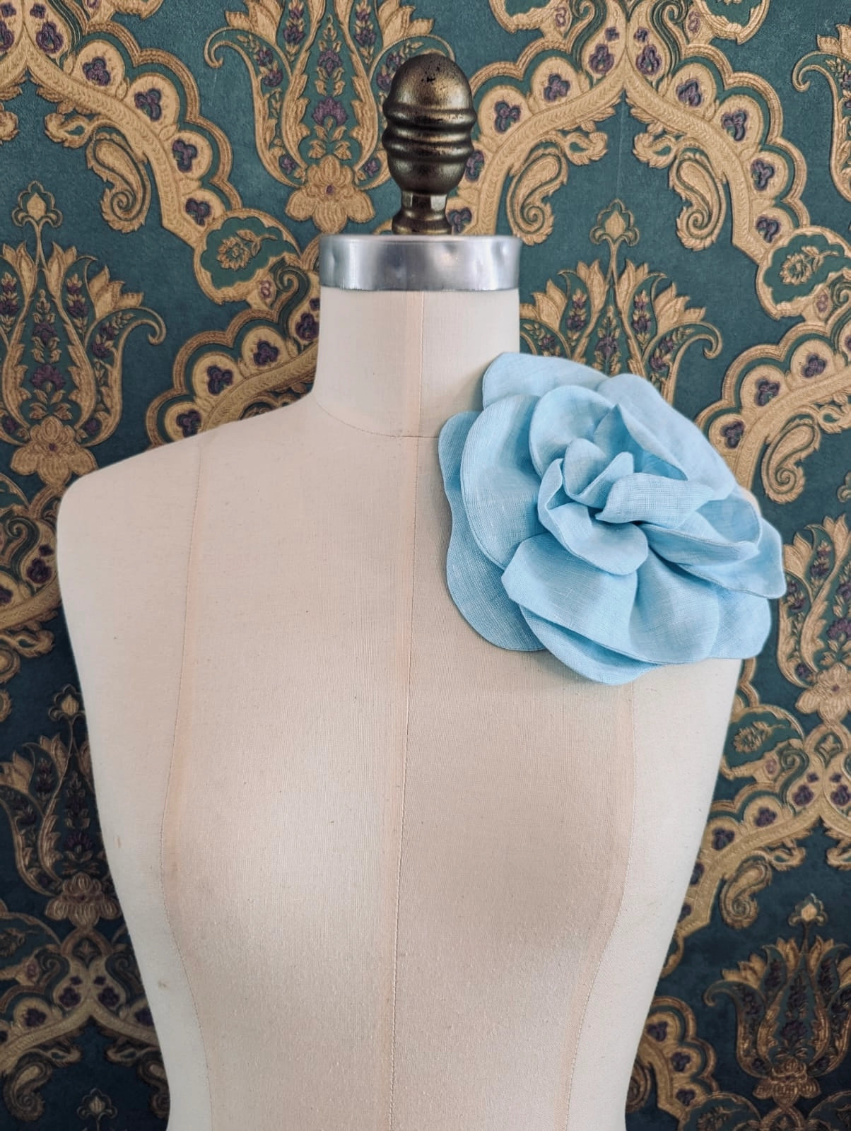 Broochella Florella large handmade linen flower brooch in blue. Pictured on a mannequin. The background is green and gold damask wallpaper.